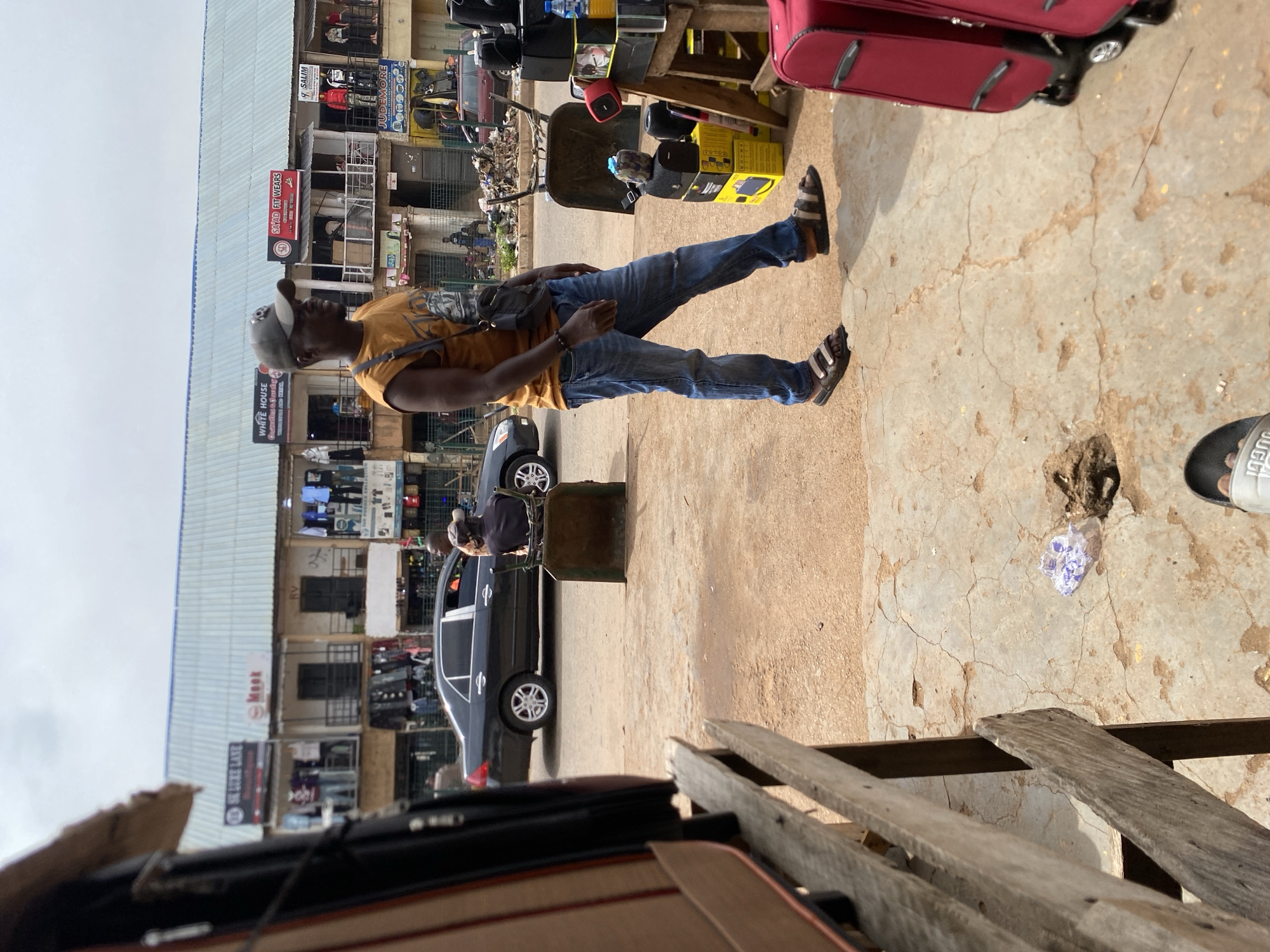 A medium shot captures a bustling street scene in Bukuru, Nigeria. The foreground features wooden structures with luggage visible, suggesting a marketplace or transportation hub. A man in an orange t-shirt and blue jeans walks towards the right, his expression neutral. He wears a dark cap and a crossbody bag. In the background, a dark sedan is parked on the road, with two men standing near it. A row of shops with signs like "SK LUKE LANE," "Meek," and "SAYAD FIT WEARS" lines the street, displaying clothing on racks. The environment is characterized by a dusty road, cracked concrete ground, and a cloudy sky. Several other individuals are faintly visible in the distance, contributing to the overall impression of a lively commercial area.