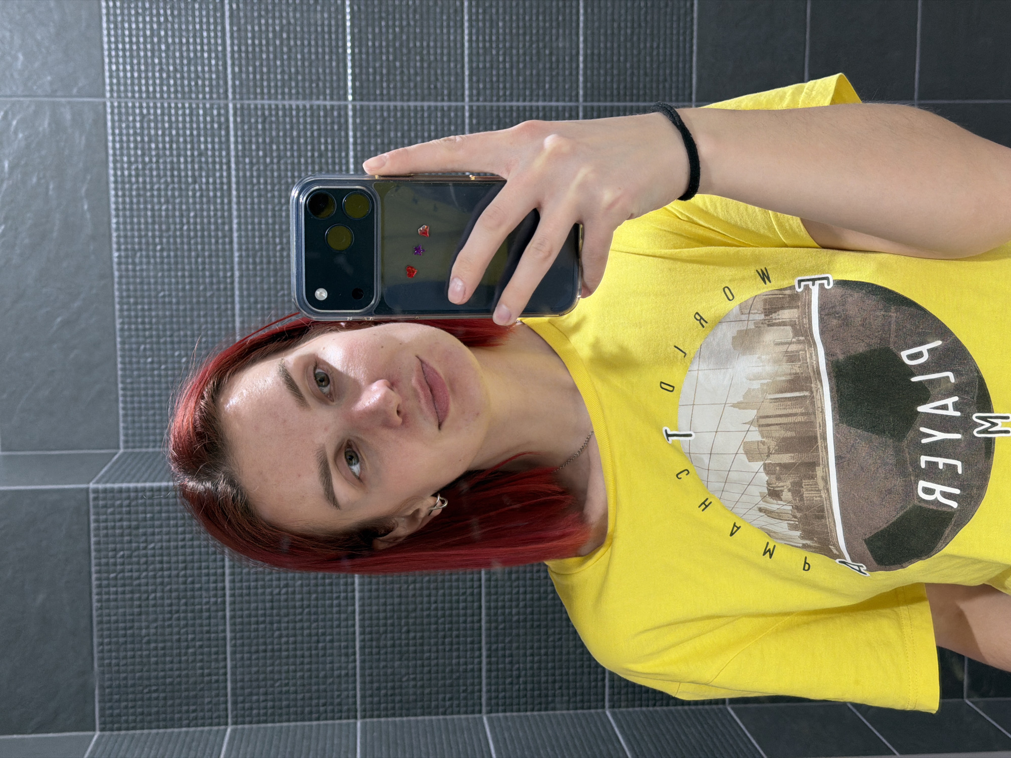 A person with red hair is taking a selfie with a smartphone in a bathroom. The background features gray tiled walls. The person is wearing a yellow t-shirt with a circular graphic and text in reverse. The phone case is clear and contains small decorative elements. The person is holding the phone with their left hand, which has a black hair tie on the wrist. The setting appears to be indoors, likely within a private residence in the city of Chaikovsky, Russia.