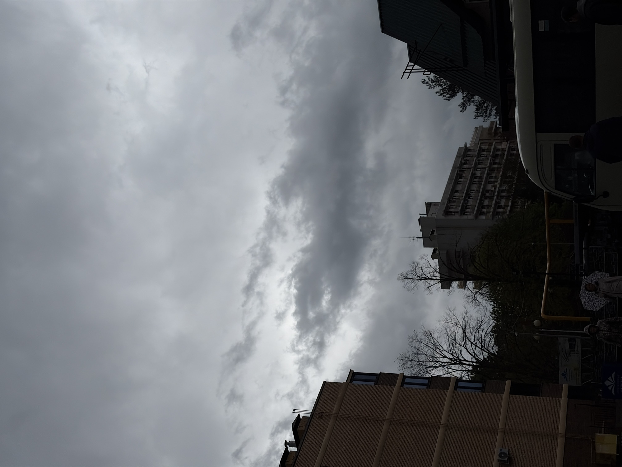 The image presents an urban street scene in Goryachiy Klyuch, Russia, dominated by a heavily overcast, grey sky. The scene is viewed upwards towards the sky, with buildings framing the composition.
The sky is filled with dense, dark grey clouds, indicating gloomy, possibly rainy weather. A particularly dark and thick band of clouds stretches across the middle of the frame (if oriented vertically), suggesting an impending downpour or ongoing light rain.
Several buildings are visible. On the right, a tall, multi-story building with numerous windows features a somewhat stark, possibly Brutalist architectural style. In the foreground, another building with a facade of light brown brick or stone shows distinct horizontal banding and several windows, with an air conditioning unit visible on its lower portion. On the far left, a structure with a dark, sloping roof is partially seen. Antennas or communication equipment are perched on some of the rooftops.
Bare, leafless trees, characteristic of late autumn or winter, are scattered among the buildings, their dark branches silhouetted against the cloudy backdrop. Some patches of green foliage are visible further in the background.
A white bus is partially visible on the right side, largely obscured by deep shadow. Near the bus, a person is seen holding an open umbrella with a distinctive circular pattern, strongly suggesting current or anticipated rain. Another person is faintly discernible further back, also partially hidden by shadows. A yellow pole, possibly part of a street sign or bus stop, stands near the bus.
The scene depicts everyday urban activity under a muted, cloudy sky, with people seemingly navigating the street, potentially waiting for public transport. The overall atmosphere is subdued due to the heavy cloud cover. No discernible text is legible in the image.