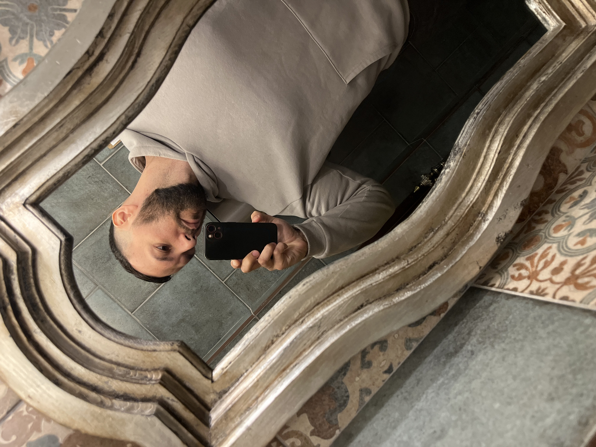 A man in a gray hoodie takes a selfie in a mirror with an ornate, antique-style frame. The reflection shows a tiled bathroom floor with contrasting patterns: gray tiles directly in front of the mirror and patterned tiles to the side. The man's face and torso are visible in the reflection, holding a black smartphone to capture the image. The location of Ulyanovsk, Russia cannot be determined from the image.