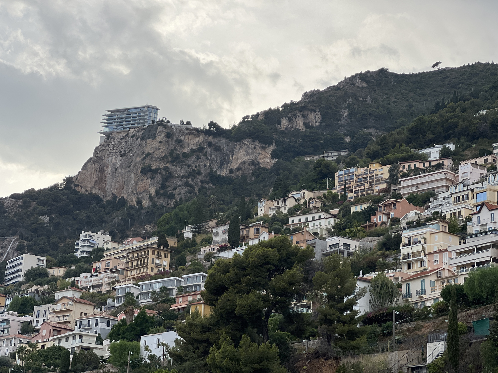 The image depicts a dense collection of multi-story buildings built into the side of a steep, rocky hill. The architecture varies, with some buildings appearing modern with glass facades, while others are older, with stucco exteriors and balconies. The buildings are clustered closely together, creating a cascading effect down the hillside. Lush green trees and vegetation are interspersed throughout the scene, softening the otherwise stark landscape of rock and concrete. The sky is overcast with grey clouds.
In the upper left of the image, a modern, multi-level building with extensive glass windows is prominently situated on a cliff edge. This building appears to be a hotel or a luxurious apartment complex. The rest of the image is filled with a dense residential area, suggesting a coastal town or city, likely Roquebrune-Cap-Martin, France, given the typical architecture and steep terrain found in this region. There are no visible people in the image, and the scene appears to be a static view of the urban landscape.