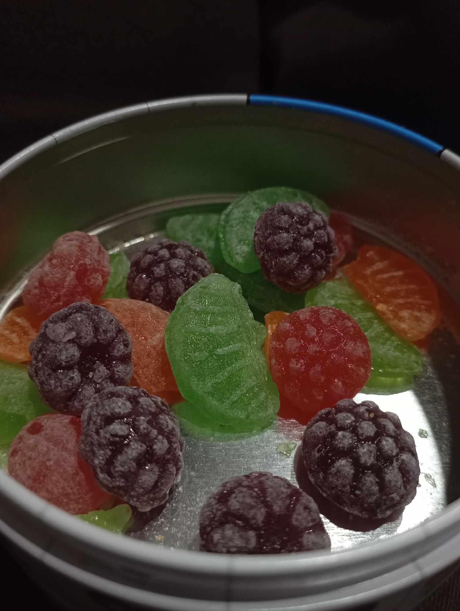 A high-angle, close-up shot shows a metal tin filled with assorted fruit-flavored gummy candies. The candies appear to be of different shapes and colors, including red, green, and orange segments shaped like citrus slices, as well as dark purple, round candies resembling blackberries or raspberries. Many of the candies have a frosty or sugary coating. The tin is silver, with a hint of blue along the rim. The background is dark and out of focus. The scene is indoors. There is no information in the image to suggest a location in Krasnodar, Russia.