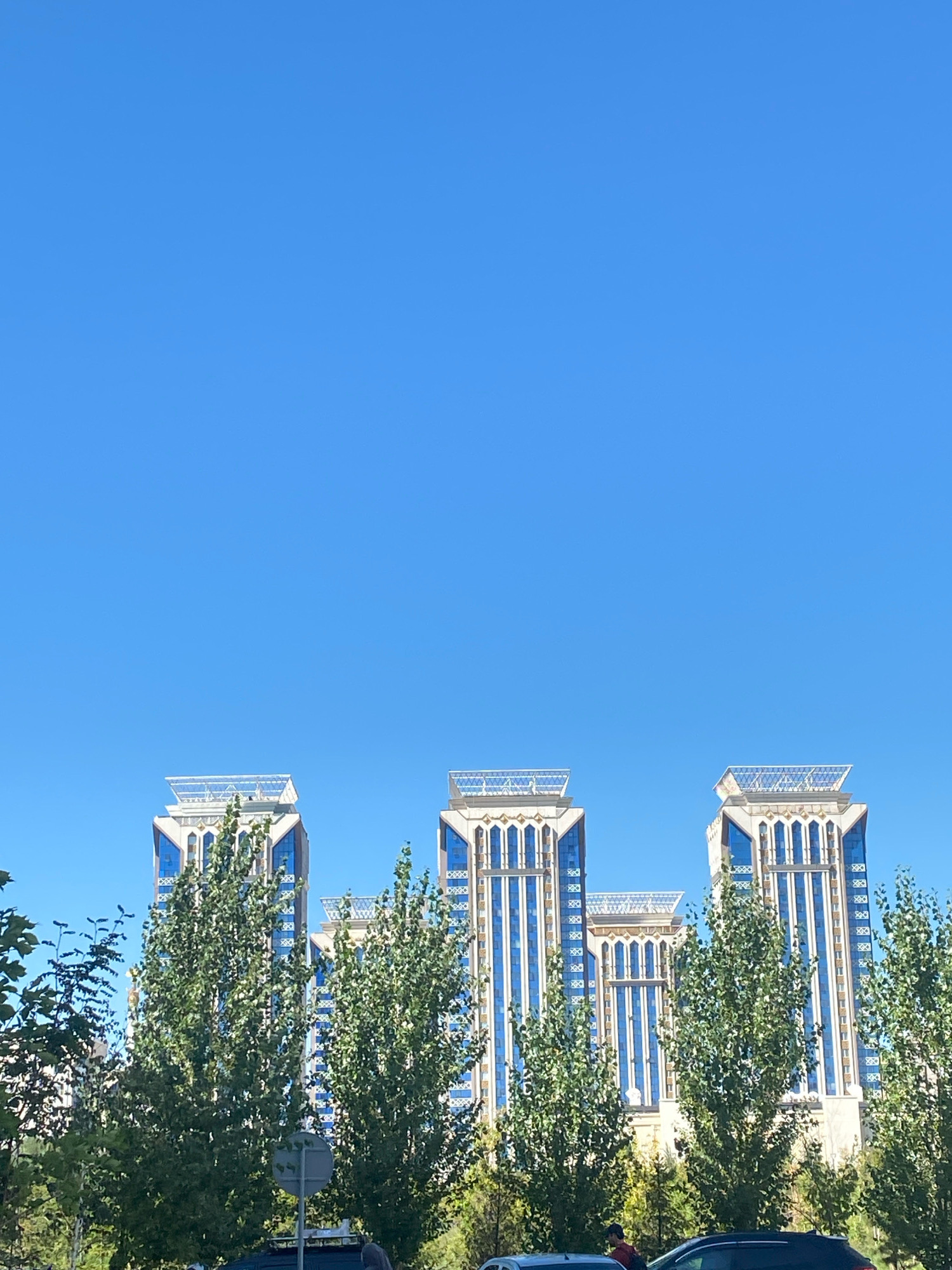 In the city of Astana, Kazakhstan, a cluster of modern skyscrapers with distinctive ornate crowns pierce a clear blue sky. The buildings feature a facade of reflective blue glass interspersed with light-colored structural elements and decorative gold accents. In the foreground, lush green trees with dense foliage partially obscure the lower sections of the buildings, suggesting a manicured urban landscape. The overall scene depicts a bright, sunny day in a contemporary city.