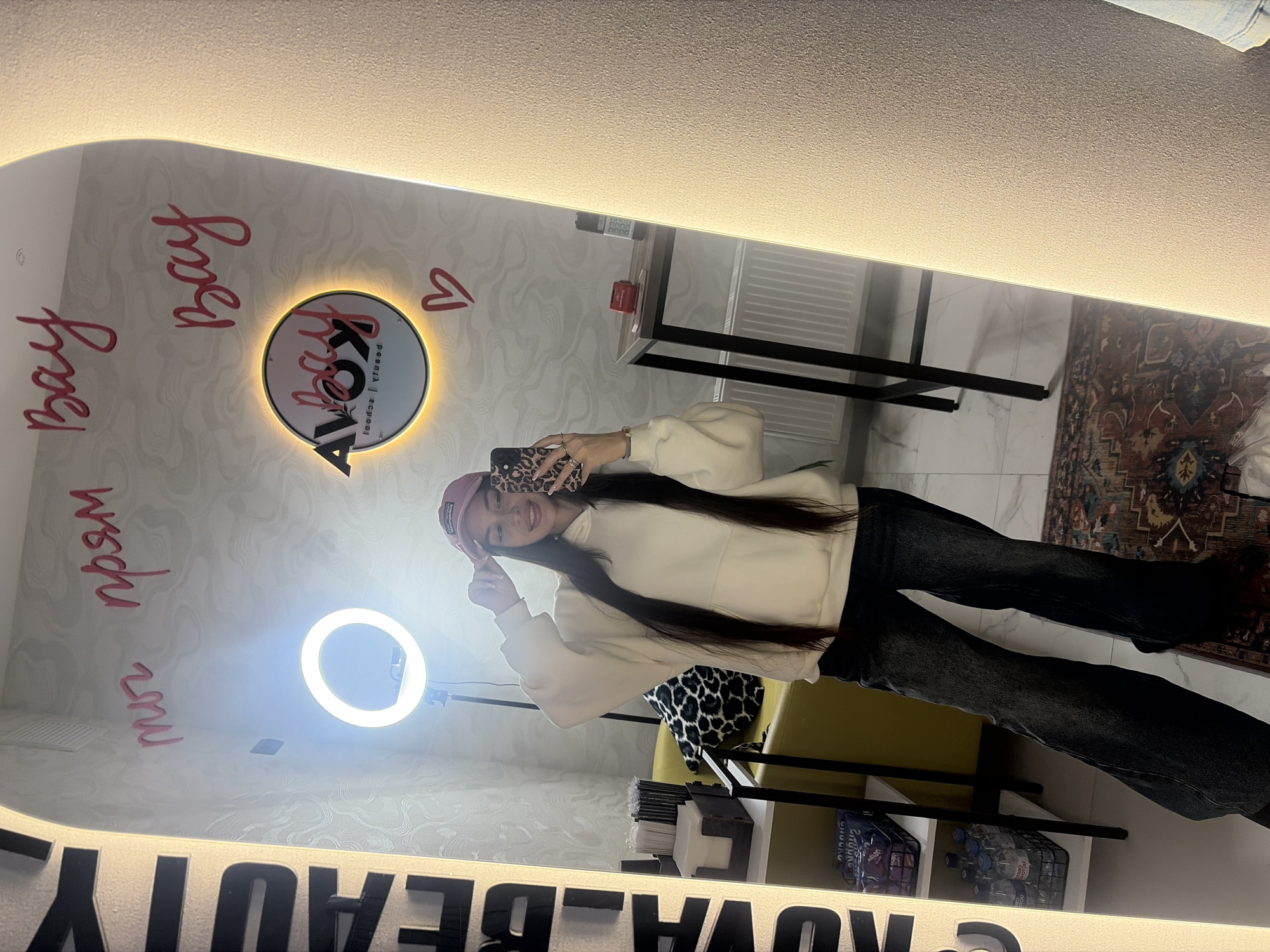 A young woman with long, dark hair, wearing a light-colored hoodie and dark jeans, is taking a mirror selfie. She is smiling and wears a pink baseball cap, holding a phone with a leopard print case. Her reflection is centered in a large, vertical, illuminated mirror.
To the left of the woman in the reflection, a bright circular ring light stands on a tripod. The background seen in the mirror reveals a modern, brightly lit interior. The walls are light-colored with a subtle wavy pattern. A circular sign with a glowing yellow halo is affixed to the wall behind her. The sign prominently features the text "KOVA" with smaller text "beauty" and "personal | school" below it. Pink script is written on the mirror surface, including "Bay Bay" (repeated) and possibly "прям мой" (Russian for "just mine"). A small red heart is also drawn on the mirror.
To the right, a black metal shelving unit holds a remote control, a small red object, and baskets containing bottled water and other items. A yellow armchair with a leopard print cushion is partially visible next to the shelving. The floor features a patterned rug over light-colored tiles. Along the bottom edge of the mirror, large black letters spell out "KOVA BEAUTY". The bright lighting, including strip lights along the mirror frame, indicates an indoor commercial setting, likely a beauty salon or studio. The woman's joyful expression suggests a pleasant activity. The location is Izhevsk, Russia.
