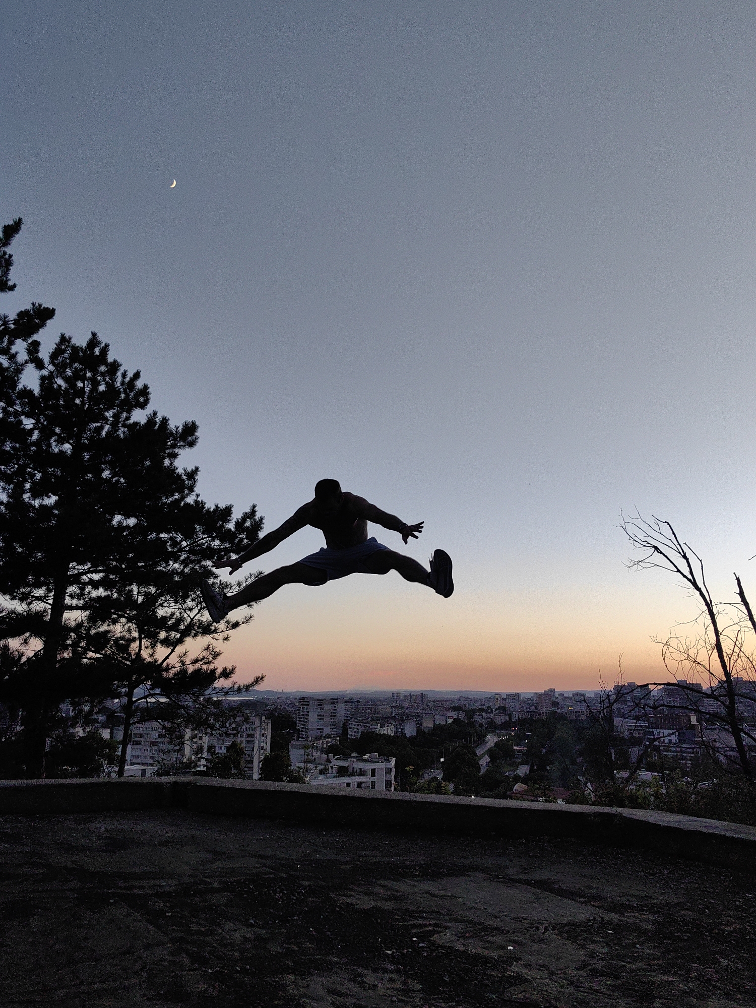 In Varna, Bulgaria, a silhouette of a man performs a wide leg jump against a twilight sky. The man is positioned centrally in the frame, suspended in mid-air with his legs extended horizontally in opposite directions, creating a dynamic split. His arms are outstretched, palms facing downwards. He is wearing shorts and athletic shoes, both appearing as dark shapes against the lighter sky.
To the left of the man, the dark silhouette of a pine tree dominates the foreground, its branches reaching upwards and partially obscuring the sky. A sliver of the crescent moon is visible in the upper left quadrant of the sky.
Below the man, the cityscape of Varna unfolds. Buildings of various heights are visible, with some appearing taller and more modern, while others seem older. The city lights are beginning to twinkle as dusk settles. The horizon is marked by a gradient of colors in the sky, transitioning from a pale blue at the top to soft oranges and pinks closer to the city. Bare, skeletal tree branches are present in the right foreground, adding a contrasting texture to the scene. The ground in the immediate foreground is dark and appears to be a rough, unpaved surface, possibly dirt or gravel.