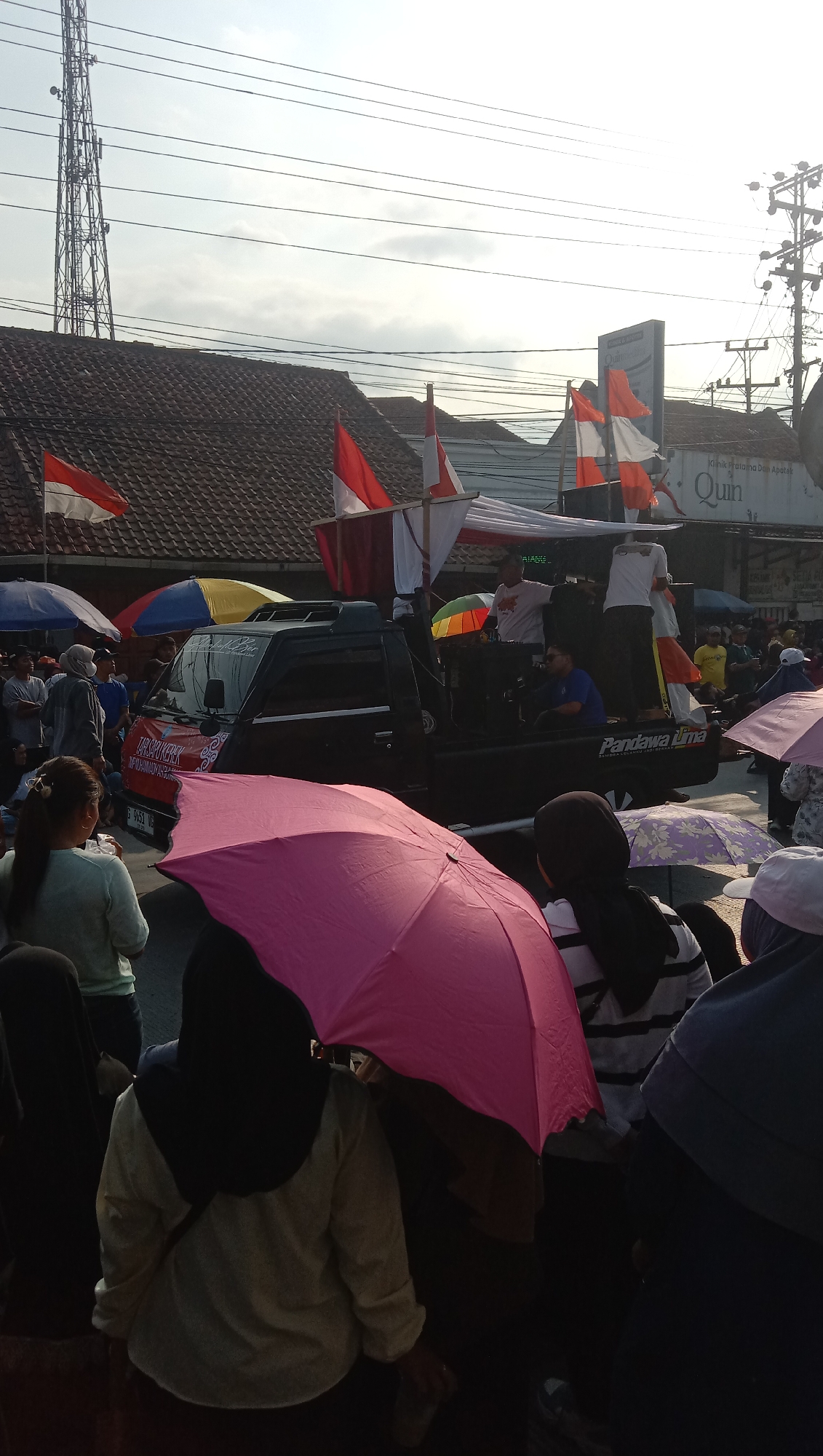 In Jatisawit, Indonesia, a group of people are gathered on a street, with many holding umbrellas. A black pickup truck is parked on the street, with several individuals standing on its platform, some appearing to be addressing the crowd. Indonesian flags are visible, some on the truck and others attached to buildings. The sky is overcast, and utility poles and wires are present in the background, indicating an urban or semi-urban setting. The overall scene suggests a public gathering or event taking place outdoors.
