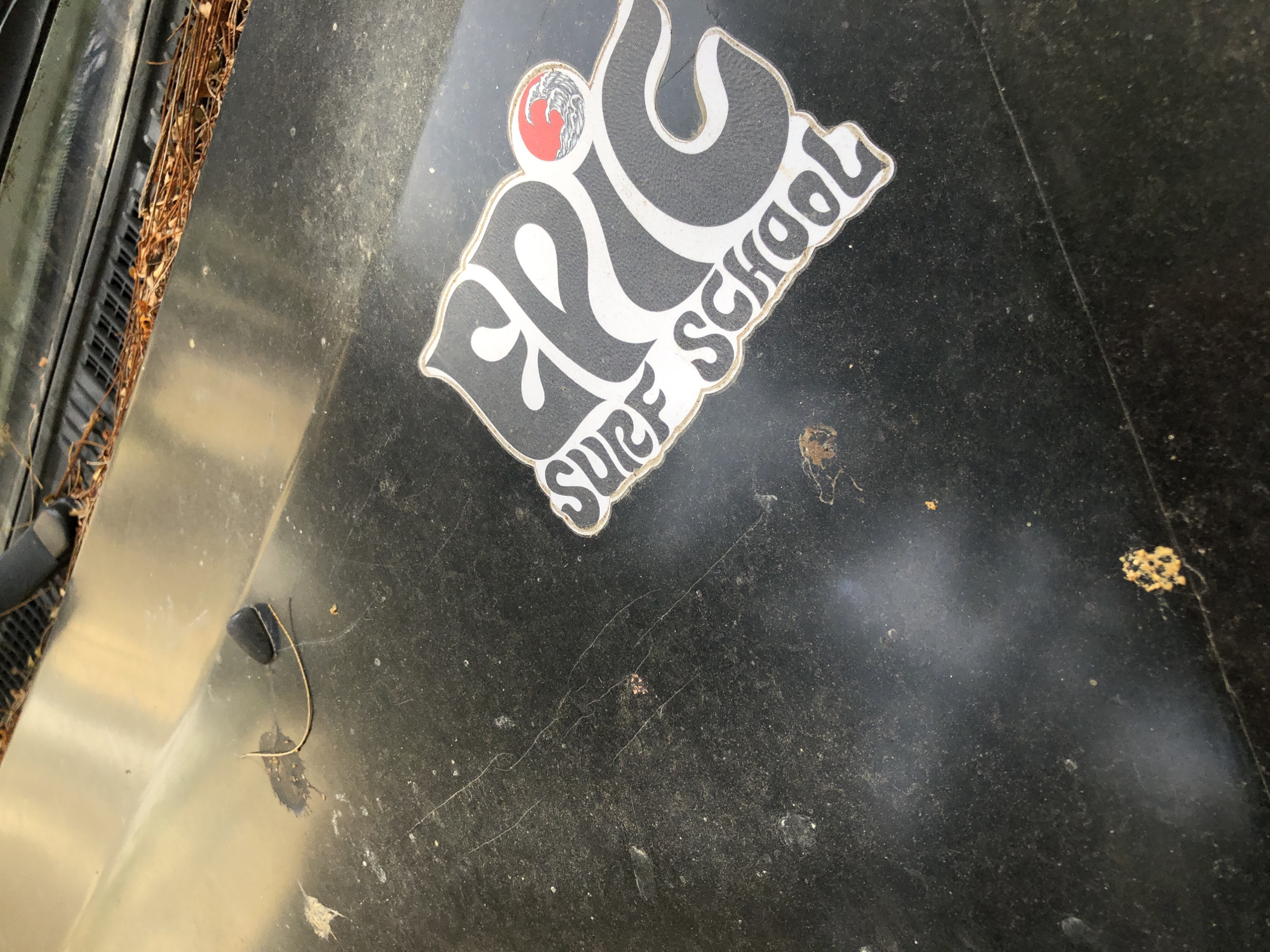 The image shows a close-up view of a dirty, black car hood with an "EPIC SURF SCHOOL" sticker. The sticker is black and white with a red sun and wave graphic in the upper right corner of the "EPIC" lettering. The hood is speckled with dust and debris, including some dried pine needles clustered near the windshield. There is a black rubber windshield wiper base and a thin, light-colored wire or string lying across the hood. The scene appears to be outdoors, possibly in a parking lot or storage area, given the dust and debris. The location is not explicitly stated, but the sticker suggests a surf school, which are common in coastal cities like Đà Nẵng, Vietnam. However, there are no specific landmarks or contextual clues in the image to confirm this location.