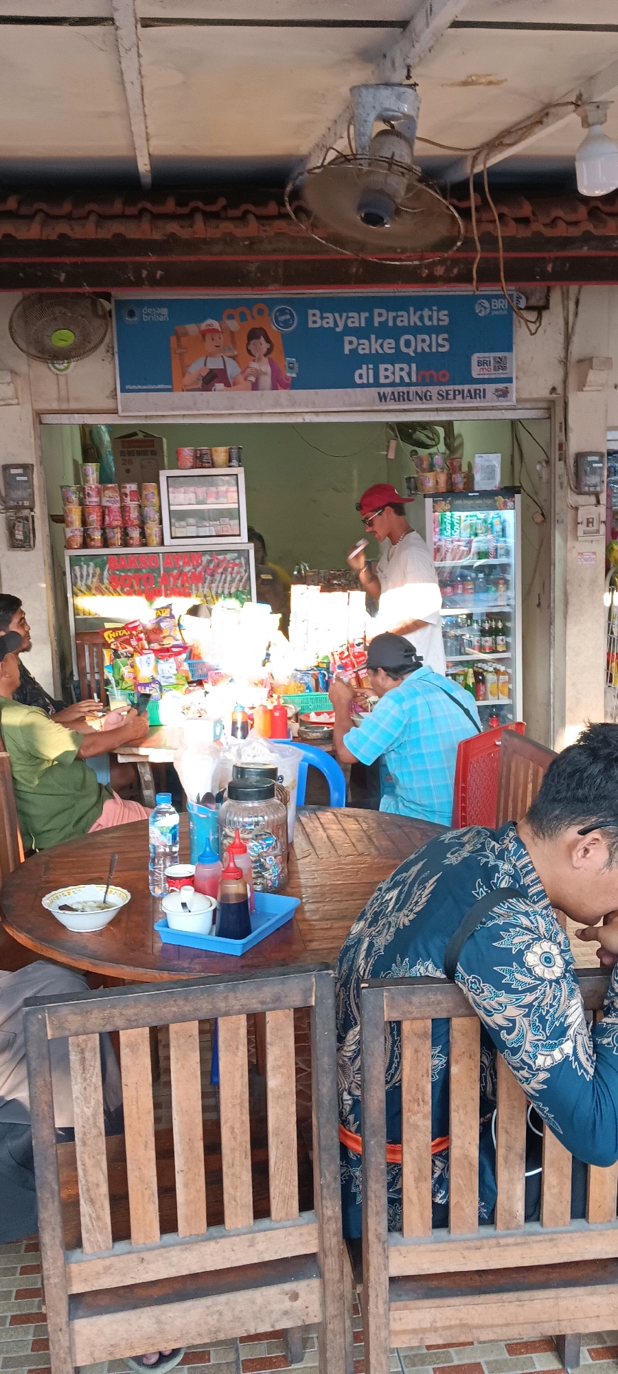 A medium shot captures a busy outdoor food stall, identified as "WARUNG SEPIARI" in Kuta Selatan, Indonesia. The scene is bustling with activity, suggesting a midday meal.
In the foreground, a round wooden table is laden with various items. A white bowl containing food, a spoon, a bottle of water, several condiment bottles (ketchup, chili sauce, soy sauce), and a small white dish are visible. Several individuals are seated around this table, with their backs partially or fully to the camera. One person, wearing a dark blue shirt with a batik pattern, is seated on the right. Another person on the left wears a green shirt.
Behind the table, the food stall itself is visible. A man in a white t-shirt and a red baseball cap is standing and appears to be serving food or interacting with customers. He is holding a white object near his mouth. To his right, a commercial refrigerator is stocked with beverages. Shelves above the refrigerator display canned goods and other items. A sign above the stall advertises "Bayar Praktis Pake QRIS di BRImo" and features cartoon illustrations of a man and a woman, promoting a payment service. Further signage indicates menu items such as "BAKSO AYAM SOTO NGANGAM".
The environment suggests a tropical setting, with an open-air structure providing shade. A ceiling fan is mounted overhead, and various electrical conduits and fixtures are visible. The flooring is tiled with a brick pattern. The overall impression is one of a local eatery serving its community.