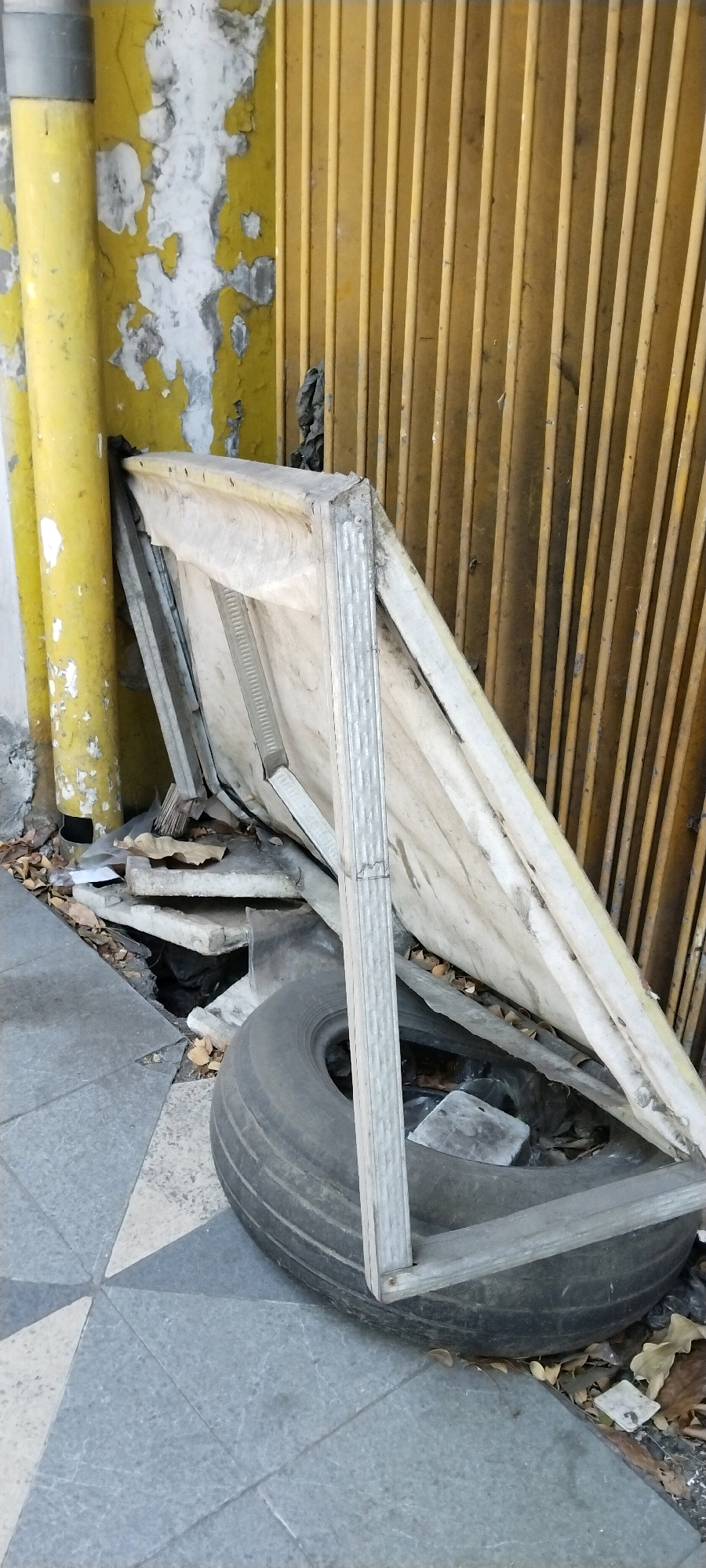 An object, possibly a discarded display stand or sign, is propped against a yellow corrugated metal gate. The object has a metal frame with a light-colored canvas or fabric covering. It is leaning on a large, flat automobile tire that rests on the ground. The ground is a mix of concrete pavement with a geometric pattern and dirt scattered with fallen leaves. To the left of the scene, a thick, yellow pipe stands vertically, its surface weathered and peeling, revealing white and gray layers underneath. The background is dominated by the vertical slats of the yellow metal gate. The overall environment suggests an outdoor urban setting, likely in a less-maintained area.