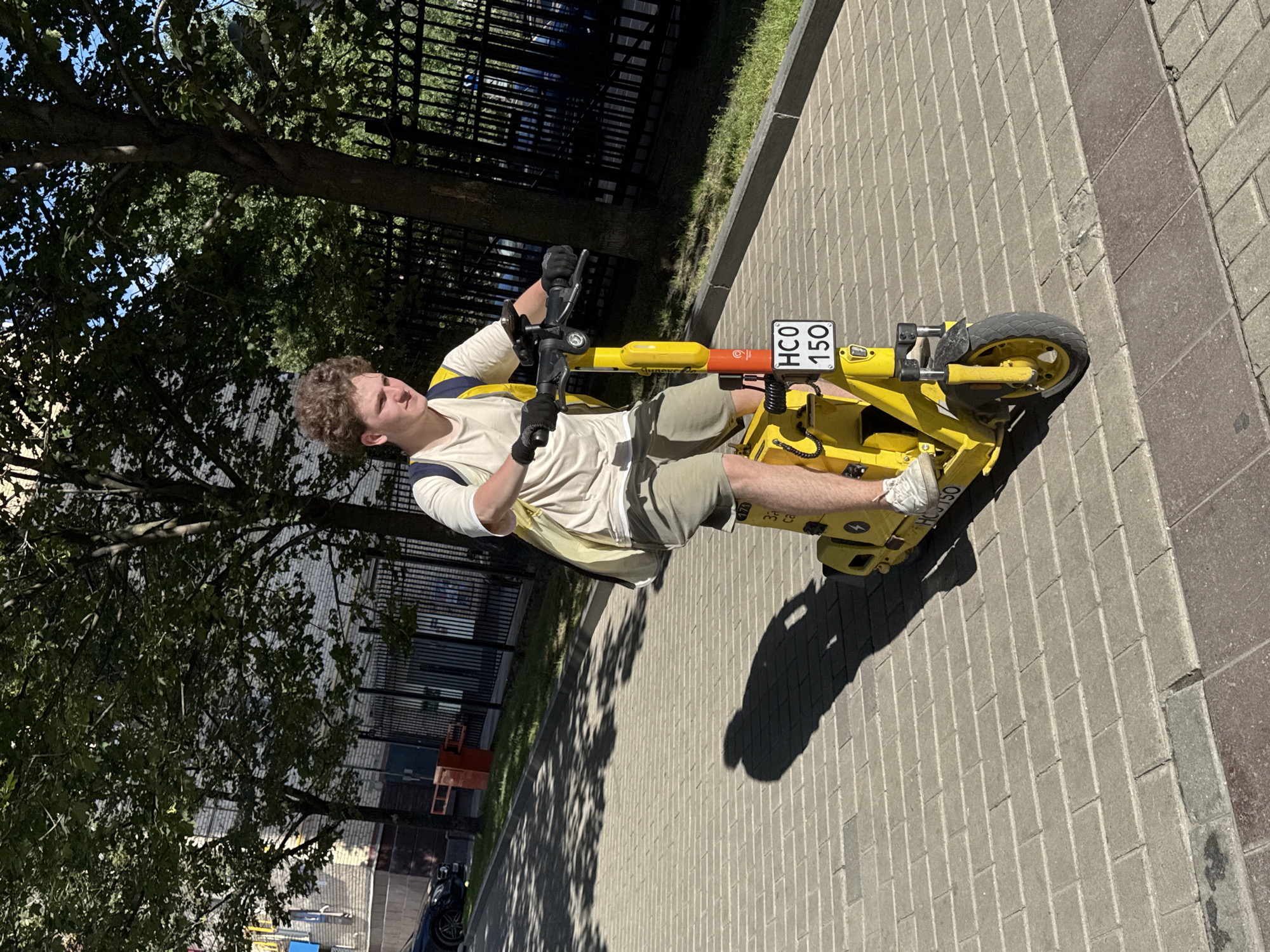 A young Caucasian male with curly brown hair is riding a yellow electric scooter on a brick-paved sidewalk in Moscow, Russia. He is wearing a light-colored t-shirt, khaki shorts, white sneakers, and black gloves. He has a yellow and blue backpack slung over his shoulders. The scooter has a license plate that reads "HCO 150". The background features a black metal fence, trees with green foliage, and a building with a brick facade. Sunlight casts shadows on the sidewalk.