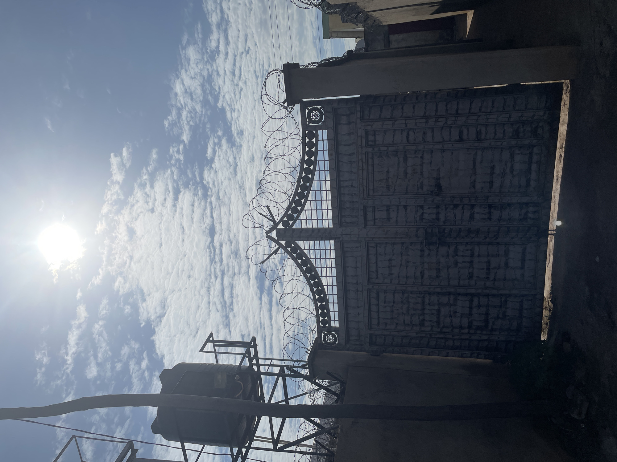 The scene captures an outdoor architectural view on a bright, sunny day in Narayi, Nigeria. A brilliant sun is visible in the upper right quadrant of a light blue sky, which is interspersed with numerous scattered white clouds. The primary subject is a large, dark, double-leaf metal gate with a textured, possibly paneled, surface, standing closed and partially obscured by deep shadow in the foreground. The gate is flanked by two substantial, light-colored concrete pillars. Above the gate, a decorative metal grille features multiple circular elements and a central rosette design. Extensive rolls of concertina wire (razor wire) are securely installed along the entire top edge of the gate and continue atop the adjacent concrete walls, extending to both the left and right out of frame, indicating robust security measures. To the far left, a large, dark plastic water tank is elevated on a sturdy metal scaffolding structure. A thick, dark vertical pole, possibly a utility pole or structural support, stands prominently near the water tank. The ground directly in front of the gate is in heavy shadow, with a narrow strip of sunlit ground visible beneath the gate's bottom edge. No individuals, animals, or active subjects are present in the image.