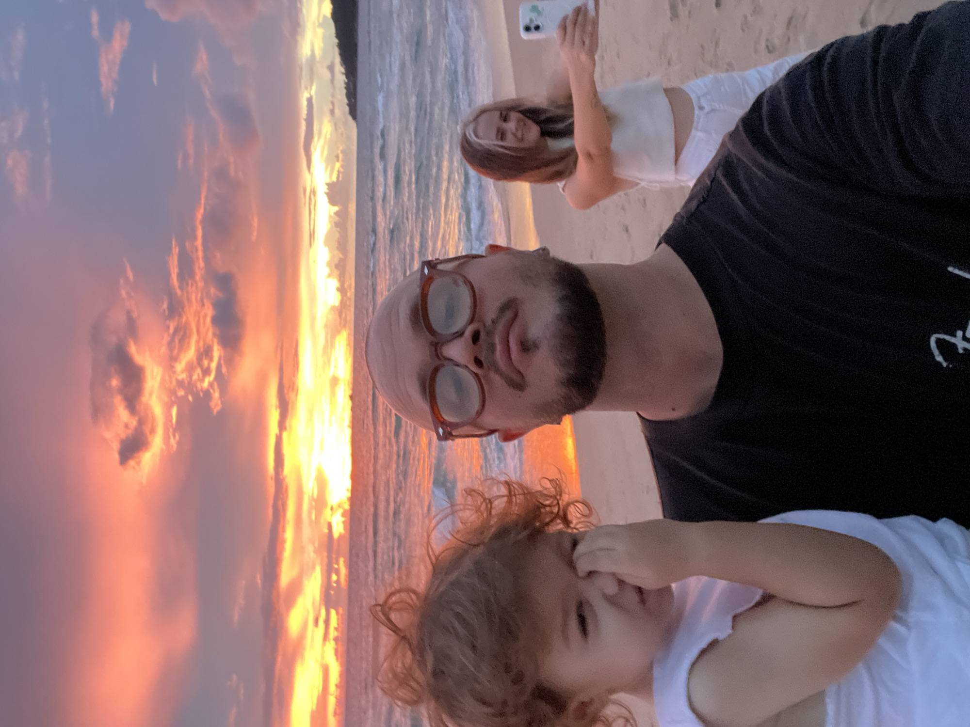 A man is holding a child in front of him on a beach at sunset. The man is bald with glasses and a beard, wearing a black t-shirt. The child, with curly red hair, is wearing a white top and rubbing their eye. In the background, a woman with blonde hair is taking a selfie with a phone. The sky is filled with vibrant orange and pink clouds from the setting sun, with the ocean waves in the middle ground and a distant shoreline. The sand is light brown.