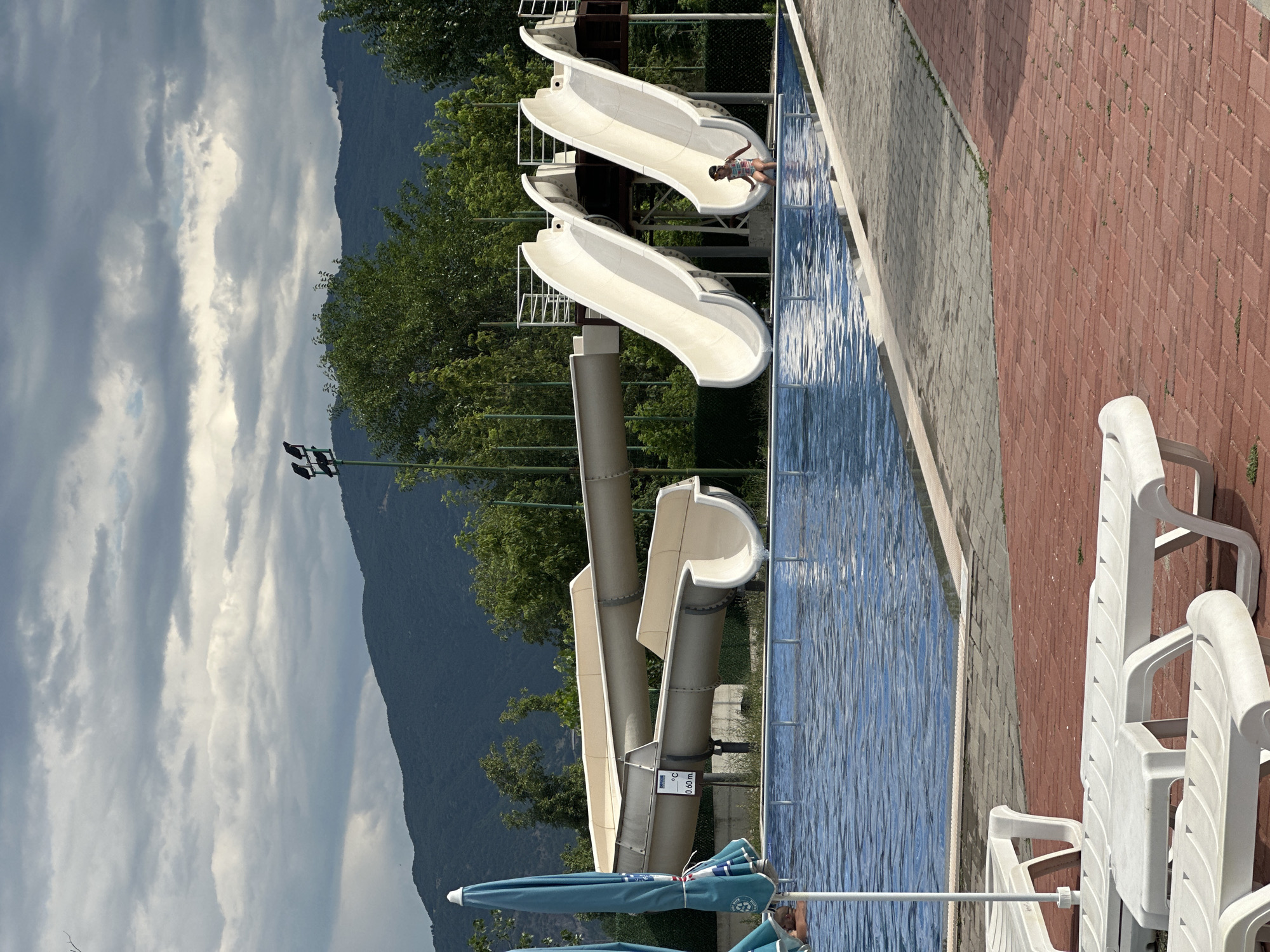 The image depicts a water park located in Simitli, Bulgaria, under a cloudy sky. In the foreground, several white plastic lounge chairs are arranged on a red brick patio. Adjacent to the patio is a swimming pool filled with blue water. Beyond the pool, a water slide complex is visible, with two large, white slides on the right and a longer, beige slide on the left. A young girl in a striped swimsuit is positioned at the bottom of one of the white slides, appearing to have just completed her ride. In the background, lush green trees and dark, forested mountains are silhouetted against the sky. A street light with multiple bulbs is mounted on a pole to the left of the slides. The overall scene captures a recreational setting at an aquatic facility.