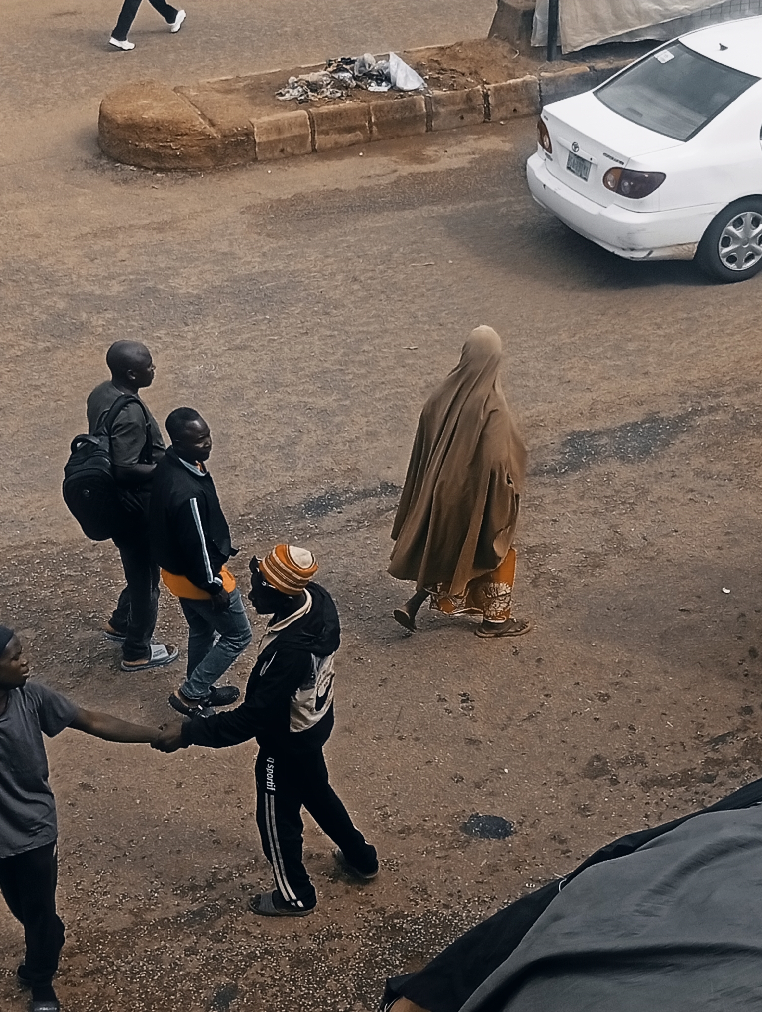 The image depicts a street scene in Bukuru, Nigeria. Several people are visible, including a person walking in the upper left corner, three men in conversation in the center of the frame, and a person in a brown hijab walking away from the camera towards the right. In the lower left, two men are shaking hands. A white Toyota sedan is parked on the right side of the image. The ground is paved with dirt and small stones. There is a curb with some debris on it in the upper part of the image. The lighting suggests it is daytime, possibly overcast. The activity appears to be casual, with people walking and interacting on a street. No text is clearly visible.