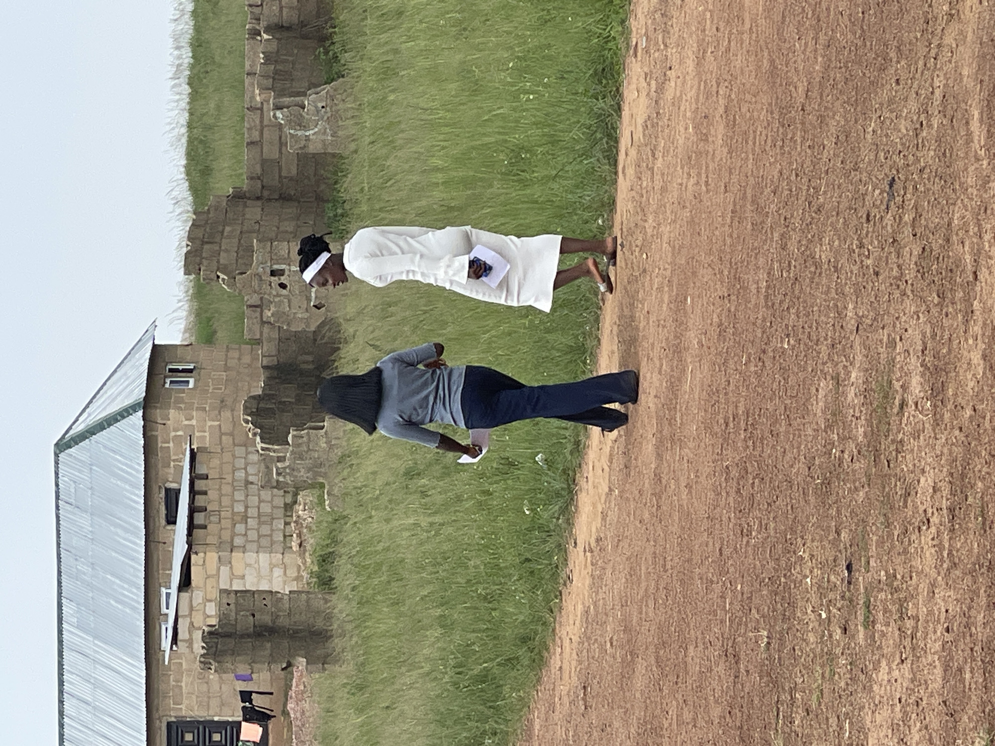 In Durumi, Nigeria, two individuals are captured in a vertical orientation. One person, dressed in white, stands with their head bowed, holding a booklet. They wear a white headband and sandals. The second person, in a grey top and dark trousers, is also looking down, holding a piece of paper. They have dark hair, partially covered by their clothing.
The scene appears to be an outdoor setting with a dirt path in the foreground and grassy, unkempt terrain in the background. To the left, a building with a corrugated metal roof and stone walls is partially visible. The architecture suggests a possibly rustic or underdeveloped area. In the distance, there are remnants of stone structures, hinting at historical or abandoned buildings.
The lighting suggests it could be daytime, but the overall mood is subdued. The people's posture and averted gazes might imply contemplation, prayer, or a moment of reflection. There is no discernible text visible in the image. The environment seems rural, with the natural elements of grass and dirt dominating the landscape around the human-made structures.