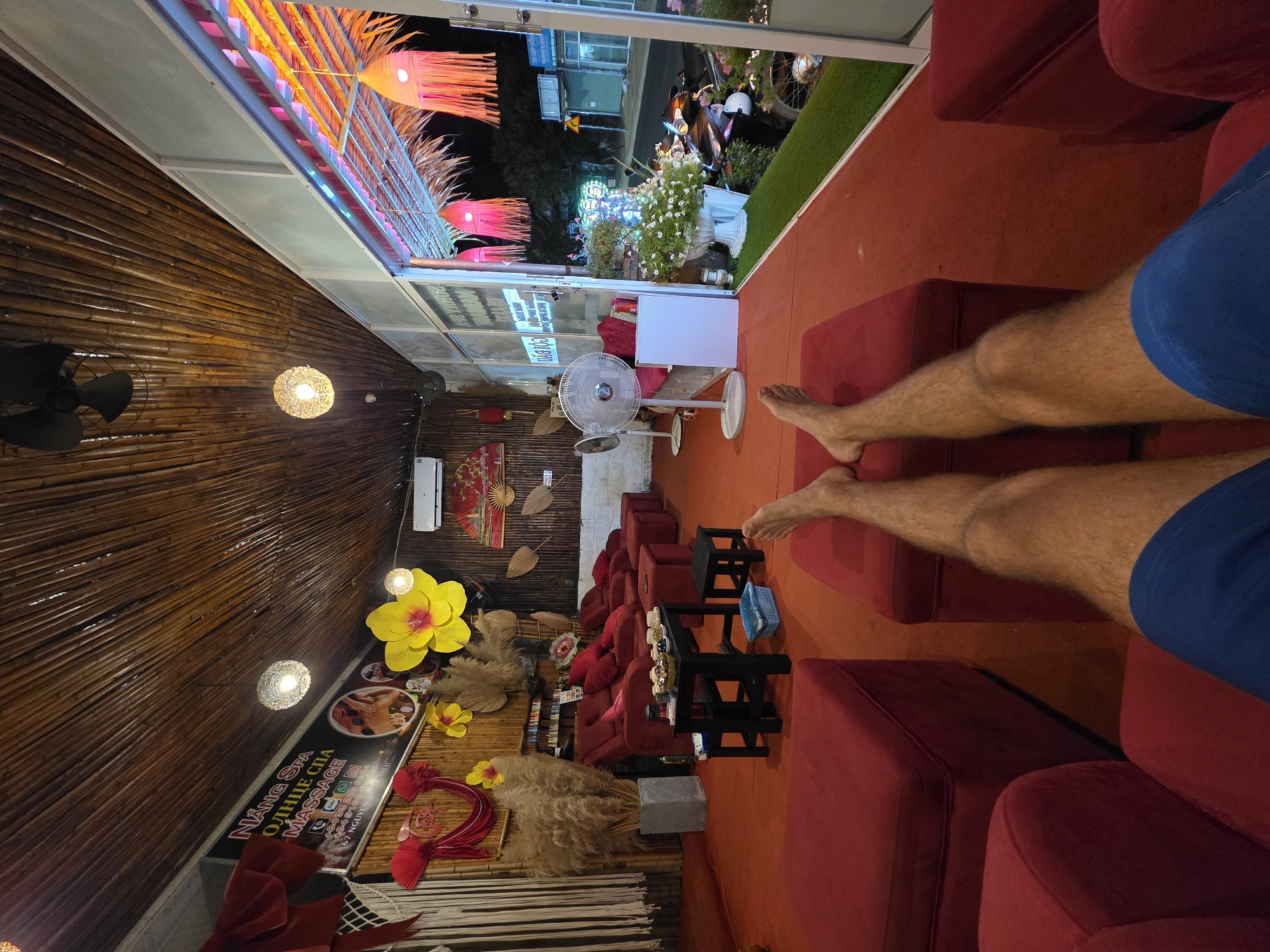 Inside a brightly lit spa in Phu Thuy Ward, Vietnam, a person in blue shorts reclines on a red cushioned seat. The spa is decorated with bamboo walls, yellow and red floral arrangements, and hanging lanterns. A sign for "NANG SPA" is visible. Outside, streetlights and passing vehicles are seen through the window. A standing fan is near the window. The floor is red.