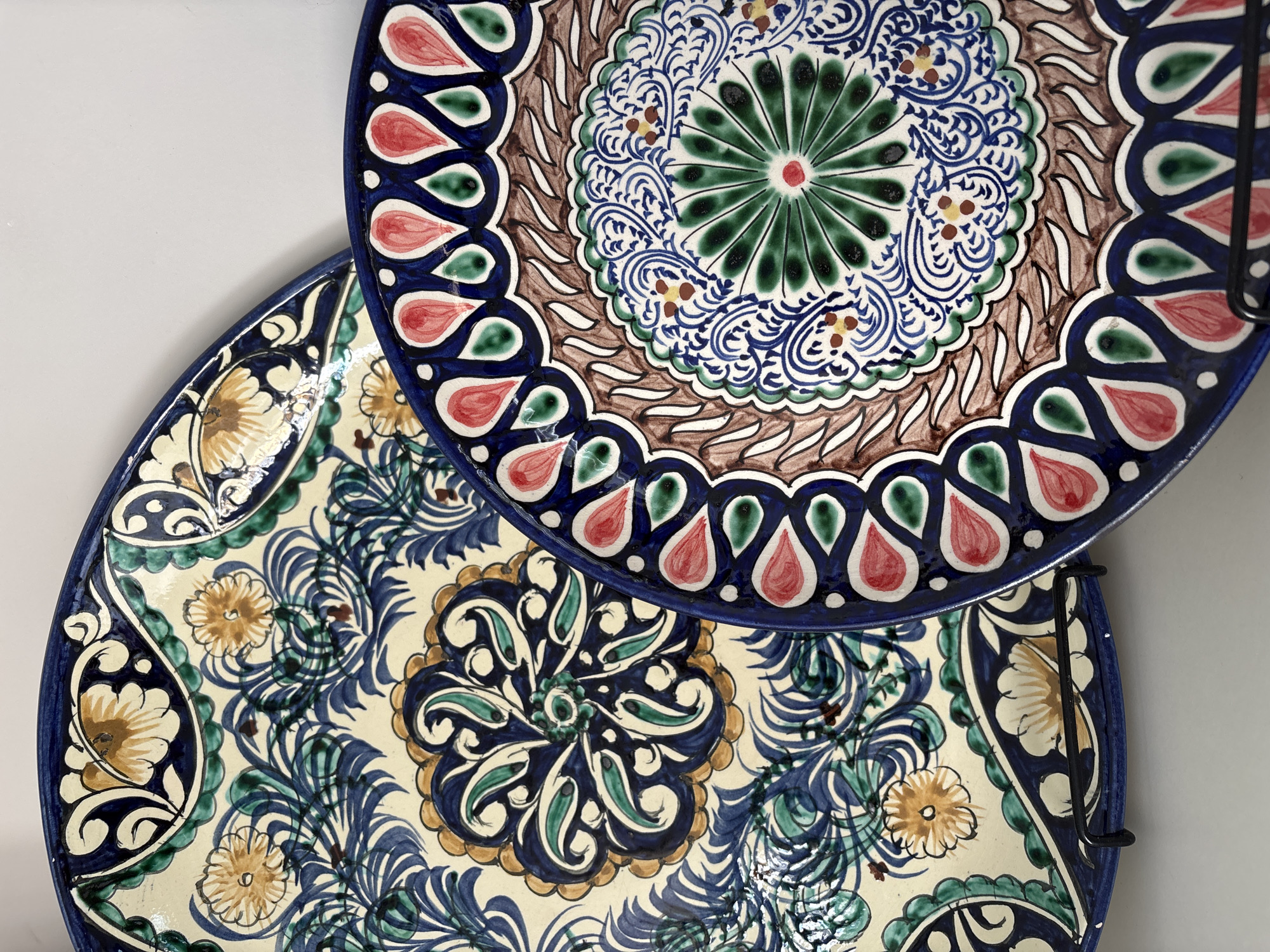 The image shows a close-up, eye-level view of two decorative ceramic plates. Both plates are round and exhibit intricate, colorful patterns characteristic of traditional Central Asian pottery. The plate on the left has a cream-colored background with a repeating floral and foliage motif in shades of blue, green, and yellow. The plate on the right is partially overlapping the first and features a more geometric central design with a starburst pattern in green and blue, bordered by red teardrop shapes and brown swirling lines. The plates are displayed against a plain, light-colored background. There is no indication of the city of Razdolnoye, Russia, in the image.