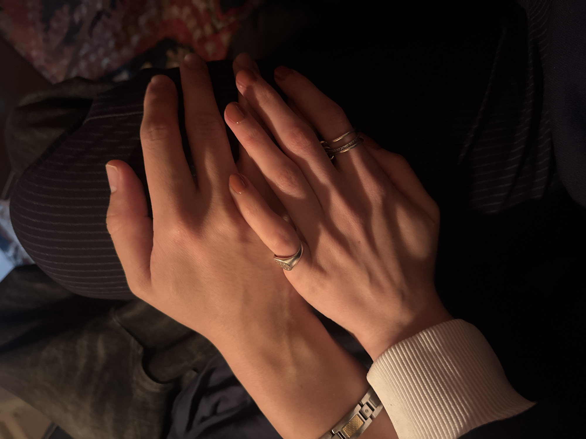 The image shows two hands resting on a lap. The hands are lightly tanned and adorned with jewelry. The right hand has two rings on the ring finger, and the left hand has a ring on the pinky finger and a watch on the wrist. The lap is covered in dark clothing. The background is out of focus, but there are hints of other objects. The image was not taken in New York City.