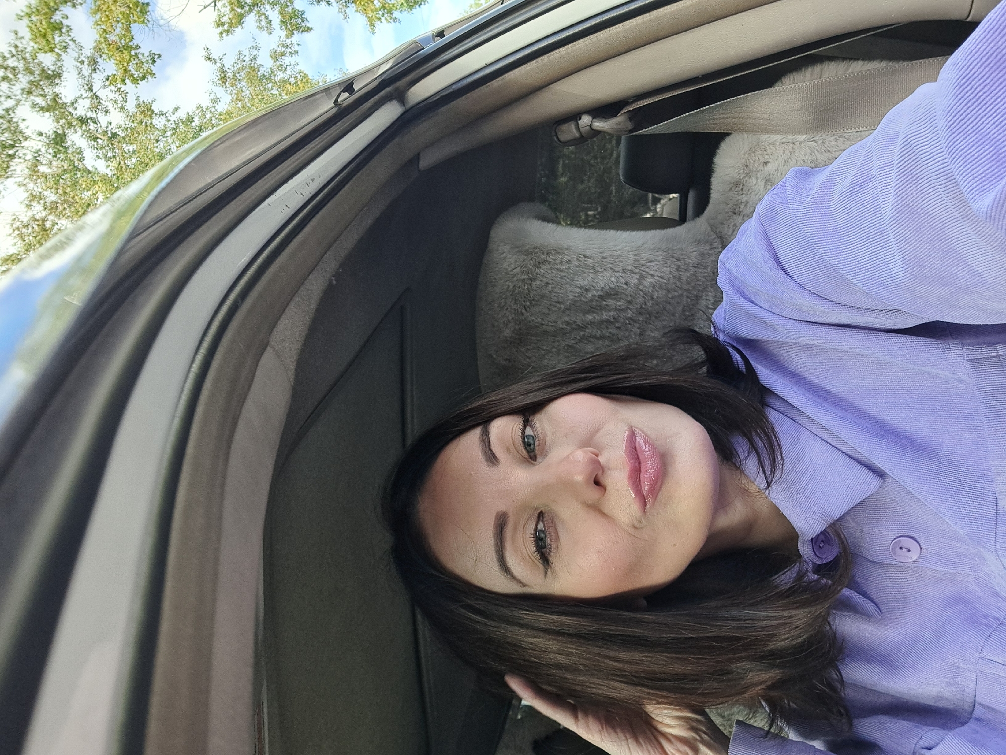 A woman is taking a selfie in the driver's seat of a car. She has dark brown hair and is wearing a light purple button-up shirt. Her lips are pursed, and her left hand is touching her hair. The car's interior is visible, including the dashboard, sunroof, and a fuzzy gray seat cover. Trees and a blue sky are visible through the car window. This image was taken in Irkutsk, Russia.
