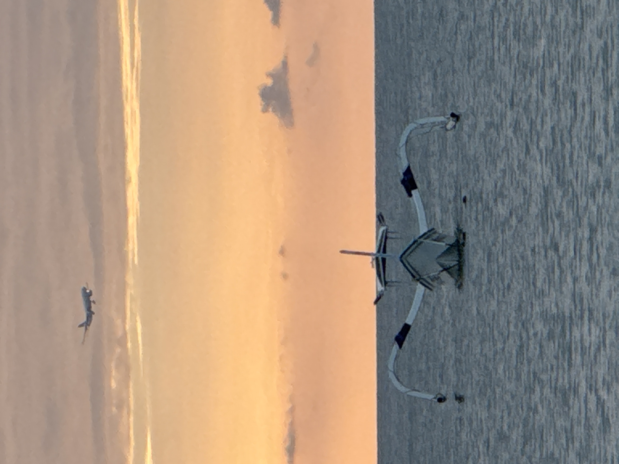 A medium shot captures a fishing boat on the water with an airplane in the sky during what appears to be sunset in Kuta Selatan, Indonesia. The sky is a gradient of peach and lavender, with wispy clouds. The airplane, a commercial jet, is visible in the upper left quadrant of the frame, appearing to be in descent with its landing gear down. The fishing boat is a traditional Balinese outrigger with a narrow hull and two outrigger booms extending to the sides. It is positioned in the center of the frame, facing the viewer. The water is calm, with gentle ripples. The overall mood is serene.