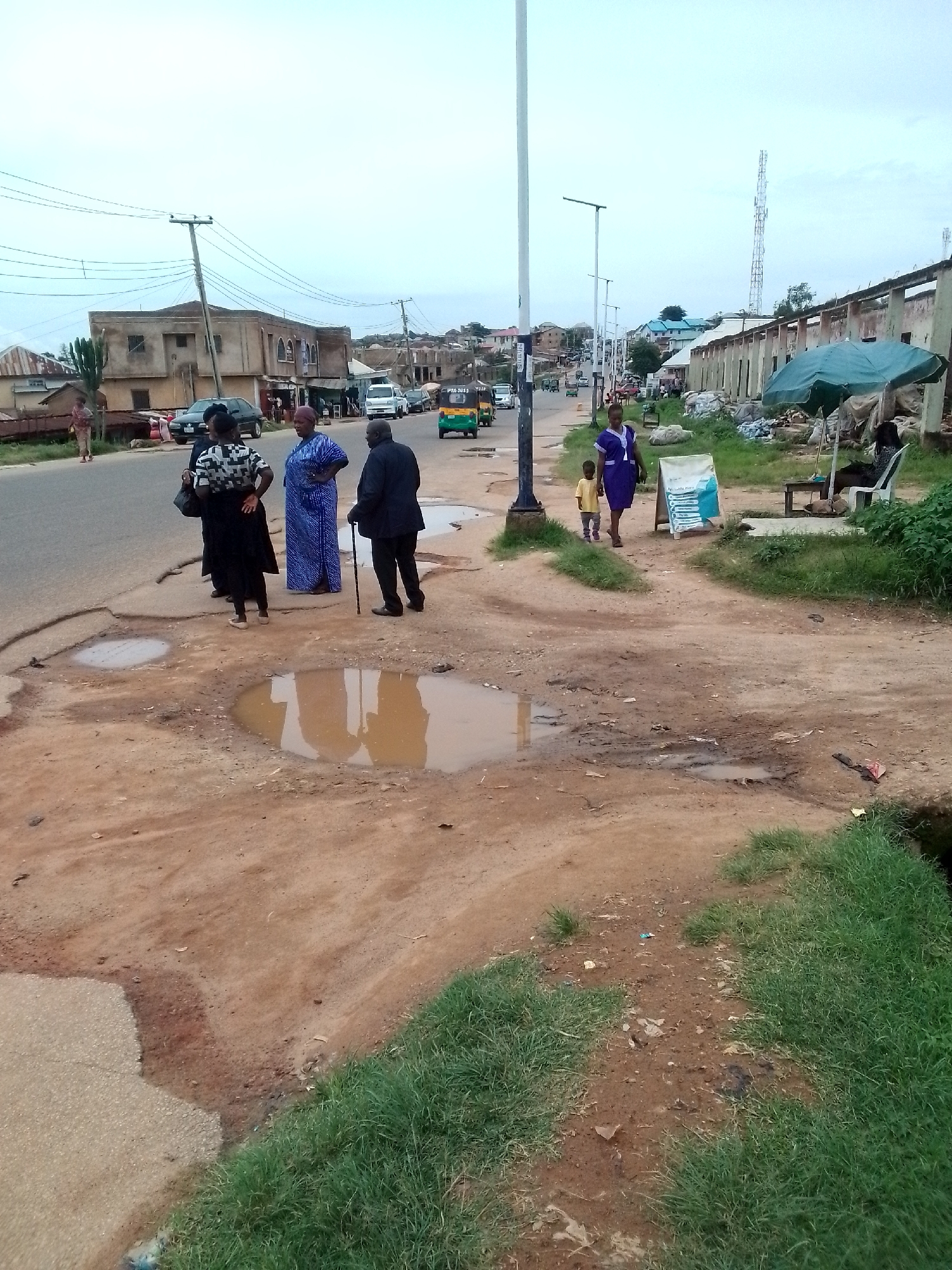 An eye-level, medium shot shows a paved road on the left, with large potholes filled with muddy water, and a dirt path with grass on the right. The sky is overcast. Several people are standing on the edge of the road, some of them appearing to be in conversation. A woman in a black dress and headscarf is facing away from the camera, with her hands on her hips. Next to her, a woman in a blue and white patterned dress is looking to her right. A man in a dark suit and hat is standing to the right of the woman in blue, holding a walking stick. To the far left, a woman in a patterned dress and headscarf stands with a young child. In the background, there are buildings, including a two-story structure with a beige facade, a white van, and a green and yellow motorized tricycle. Further down the road, a tall cell tower is visible, along with more buildings and trees. The dirt path on the right has some litter and a green umbrella providing shade over a seated person.
