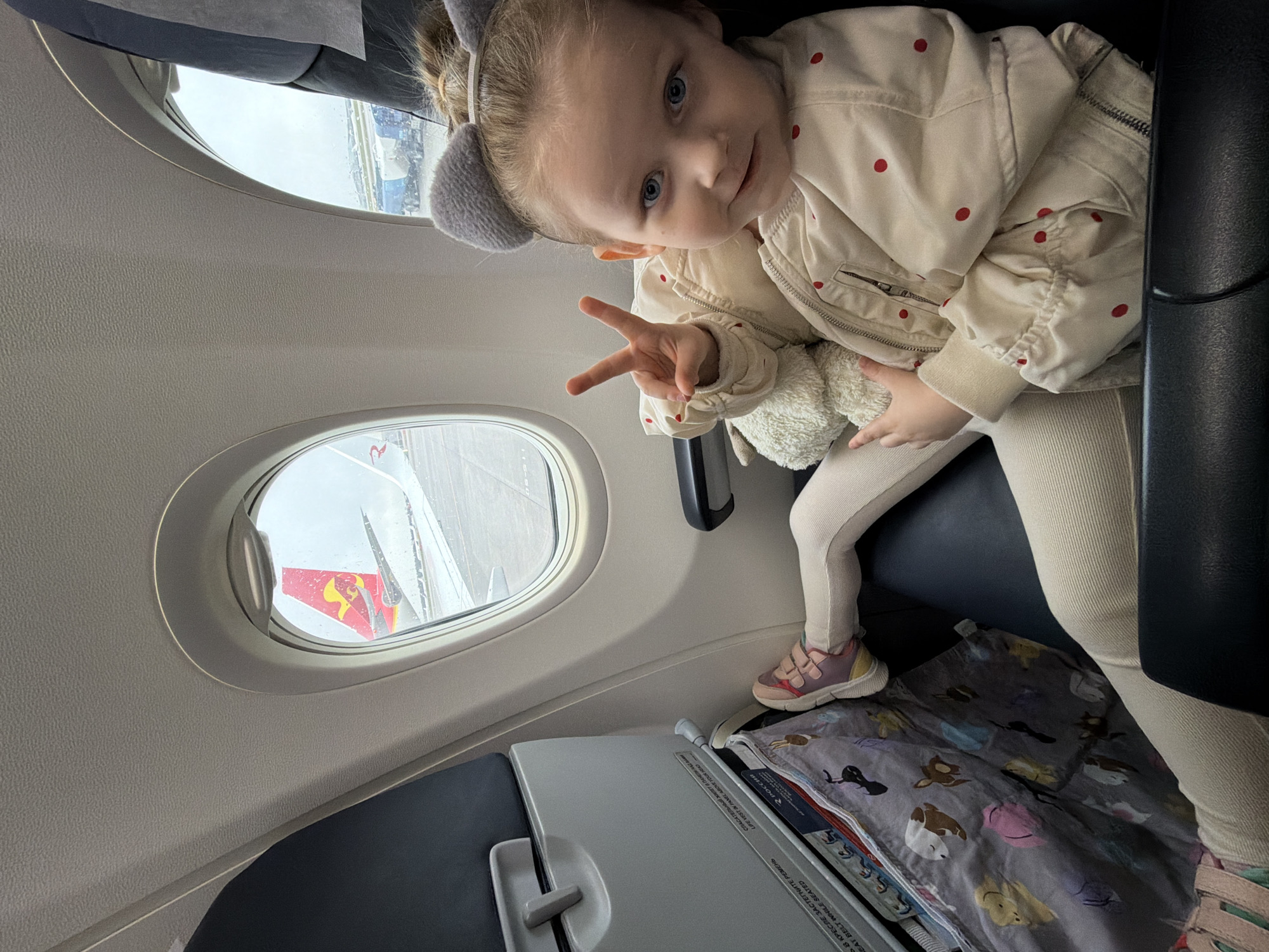A young child with light skin, wearing a cream-colored jacket with red polka dots and matching leggings, sits in an airplane seat. The child has blue eyes and light-colored hair with grey ear-shaped accessories. The child is holding a stuffed animal and gesturing with their left hand. Outside the airplane window, the wing of the plane is visible, and the airport appears to be wet. Below the child, a tray table is down and covered with a blanket featuring cartoon animal designs. The safety instructions label on the table shows Cyrillic script, suggesting the airplane is a Russian one. The image was probably taken in Khimki, Russia.