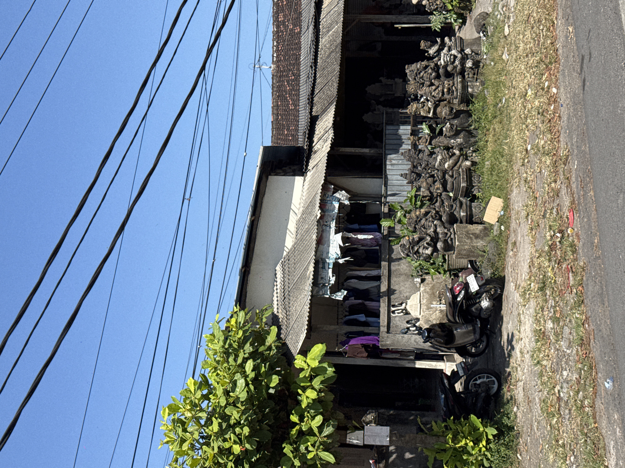 The image depicts a roadside scene in Sukawati, Indonesia, featuring a small shop or stall selling carved stone statues. The shop has a corrugated metal roof extending over an open-air display area. In front of the shop, a dense collection of intricately carved stone statues, many appearing to be of mythical creatures or deities, are arranged on the ground. Some of these statues are on pedestals. A motorcycle is parked to the left of the statues, and another motorcycle is parked in front of the shop. Above the shop, laundry hangs on a clothesline. A large, leafy green tree with bright green foliage is positioned to the left of the frame, partially obscuring the shop's entrance. Power lines are visible crisscrossing the bright blue sky overhead. The ground in front of the shop is a mixture of dirt, gravel, and sparse dry grass, with some litter visible on the roadside. The overall impression is a typical commercial streetscape in a tropical location.