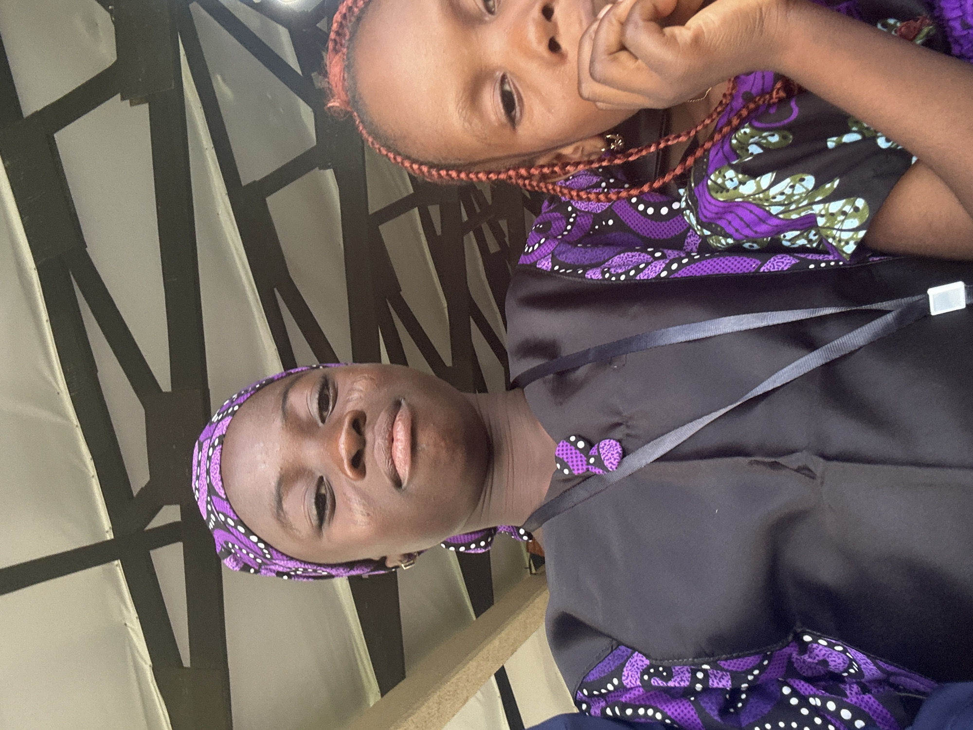 Two young girls are posing for a photo under a covered outdoor area. The girl in the foreground on the left is wearing a purple and white patterned headscarf and a black top with purple trim on the sleeves and a decorative purple button. She is looking directly at the camera with a neutral expression. To her right, a second girl is visible, wearing a similar patterned top. Her hair is styled in braids with reddish highlights, and she is bringing her hand to her mouth. The background consists of a white roof with dark beams, suggesting an open-air pavilion or canopy. The setting appears to be Itoga, Nigeria.