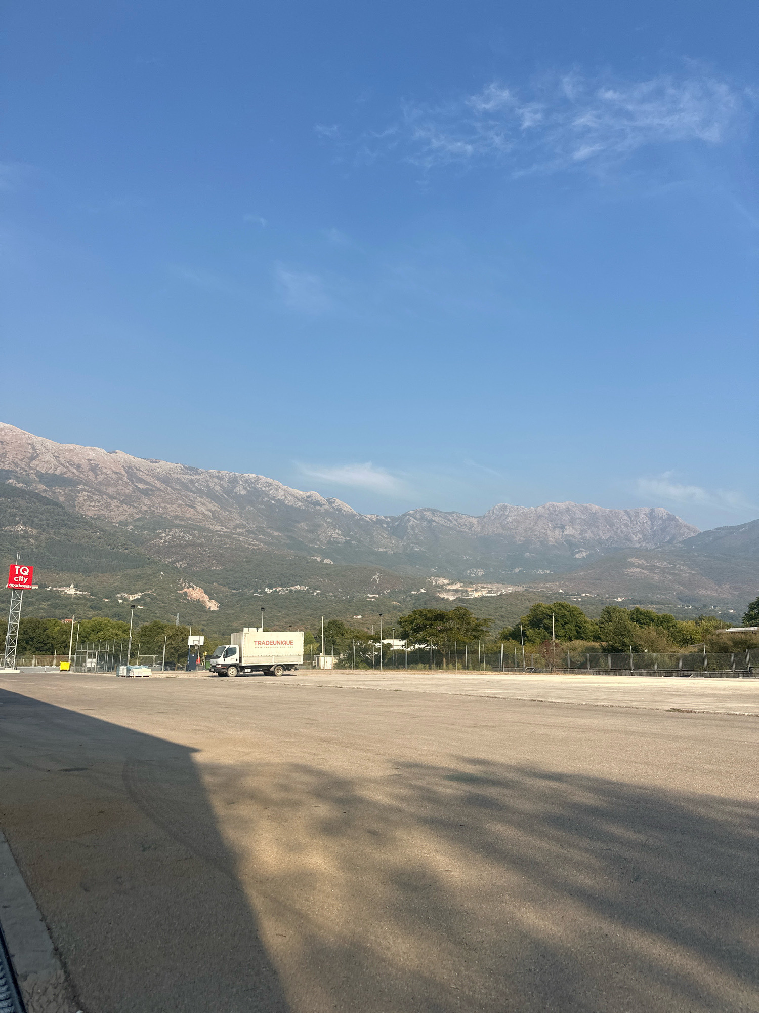 In Radanovići, Montenegro, an open, paved lot is depicted under a clear blue sky with scattered wispy clouds. In the distance, a range of rugged, sun-drenched mountains rises above a verdant, treed hillside. A white box truck with "TRADEUNIQUE" emblazoned on its side is parked in the middle ground. To the left, a red sign with white lettering reads "TQ City apartments." Fencing and scattered trees mark the transition between the lot and the natural landscape. Shadows stretch across the paved surface, indicating the direction of sunlight.
