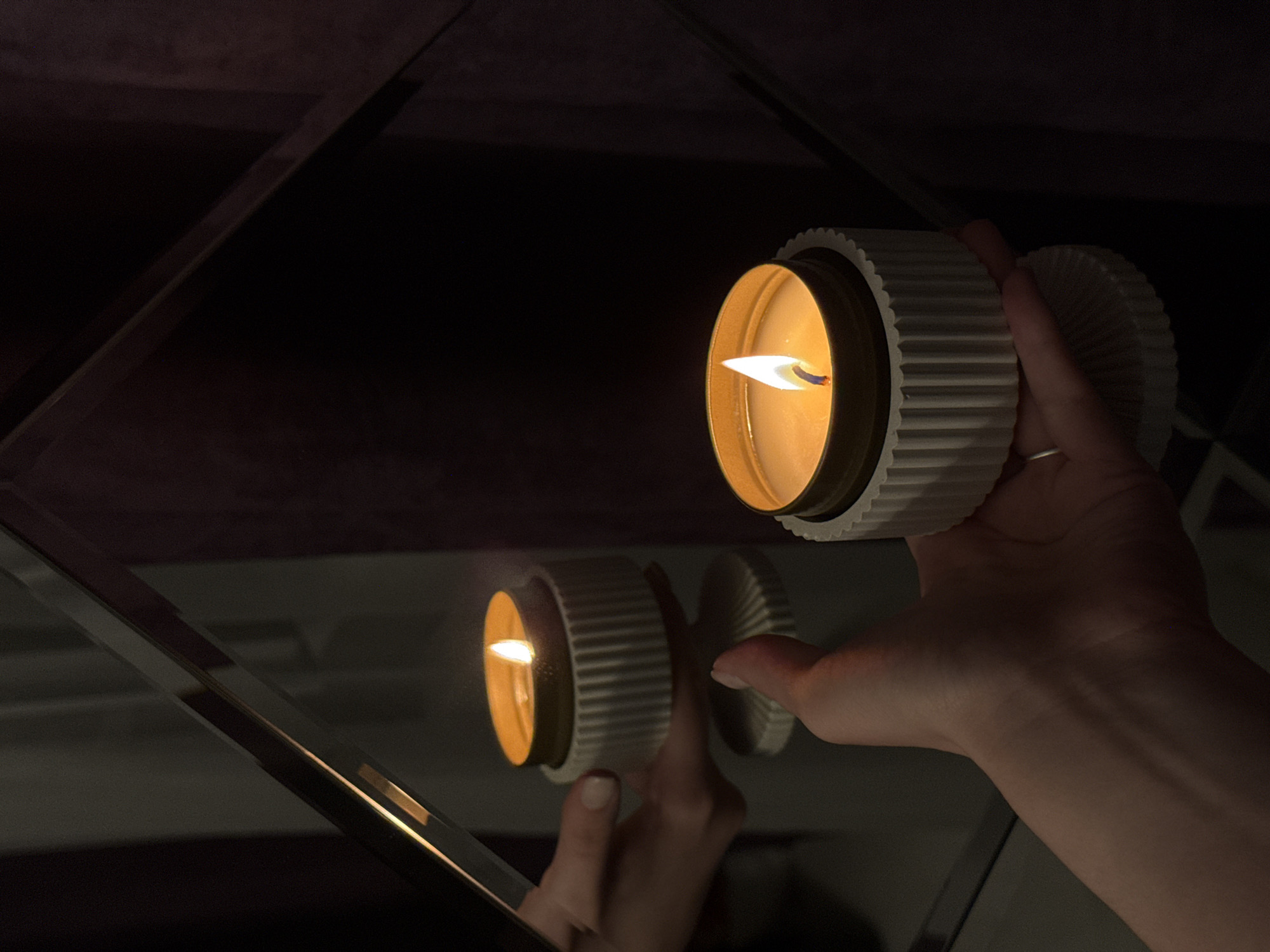 A close-up shot features a lit candle held in a person's hand. The candle has a white, corrugated outer casing and a golden interior that holds the wax and wick. The flame is small but distinctly visible. The candle is being held up to a mirrored surface, creating a clear reflection of the candle and the hand holding it. The background is dark and out of focus, suggesting an indoor setting with low lighting. The mirrored surface is made up of angled panes, creating a fragmented reflection. The lighting in the scene is dominated by the candle's glow, which highlights the textures of the candle and the details of the hand.