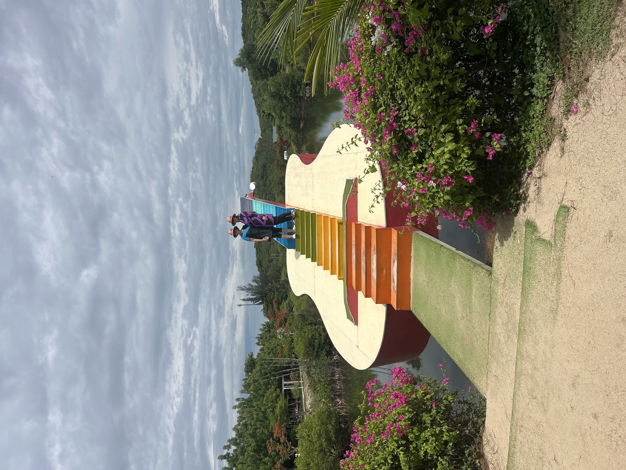 This is a full outdoor, eye-level shot of the guitar-shaped bridge in Hoa Thang, Vietnam. The bridge is a vibrant, rainbow-colored structure that spans a body of water. Two individuals are standing on the bridge, posing for a photo. The person on the left is wearing a blue shirt and a straw hat, while the person on the right is wearing a purple and black patterned outfit and a straw hat. The bridge is surrounded by lush greenery, including trees and bushes with pink and white flowers. The sky is overcast with clouds. The water beneath the bridge is dark and reflects the colors of the bridge. The foreground of the image shows a sandy area with some grass.