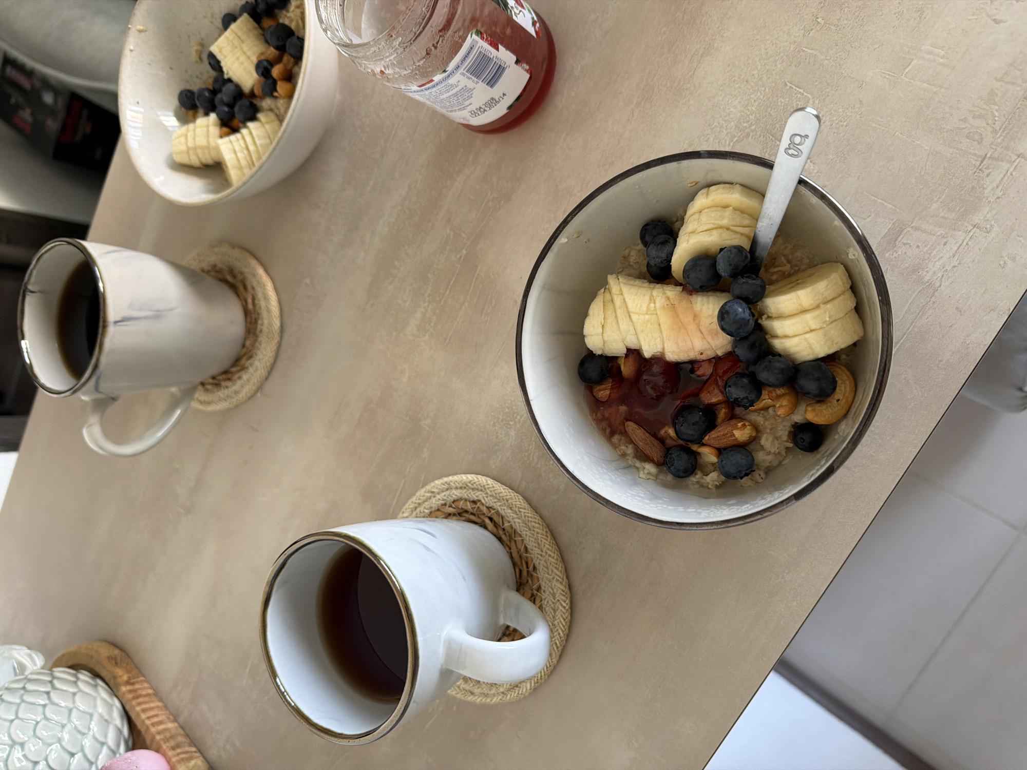 This is a high-angle, medium shot of a breakfast scene on a light-colored, textured tabletop. In the foreground, a white bowl with a dark rim contains oatmeal topped with sliced bananas, blueberries, almonds, cashews, and a red jam or sauce. A silver spoon with an etched design rests partially in the bowl. To the left of the bowl is a white mug with a marbled gray pattern, filled with dark liquid, sitting on a woven coaster. Further to the left and slightly out of focus is a decorative object on a wooden tray. To the right of the oatmeal bowl is a partially open jar of red jam with a label visible. In the background, another white mug with a marbled pattern, filled with dark liquid, sits on a similar woven coaster, positioned behind the oatmeal bowl. The setting appears to be an indoor environment, possibly a home or cafe, with a blurred background that includes a chair and some items on a shelf. There are no people visible in the image. The overall impression is a well-lit, close-up view of a healthy breakfast. This scene is not specifically indicative of Kyiv, Ukraine, as it is a common breakfast setup that could be found anywhere.