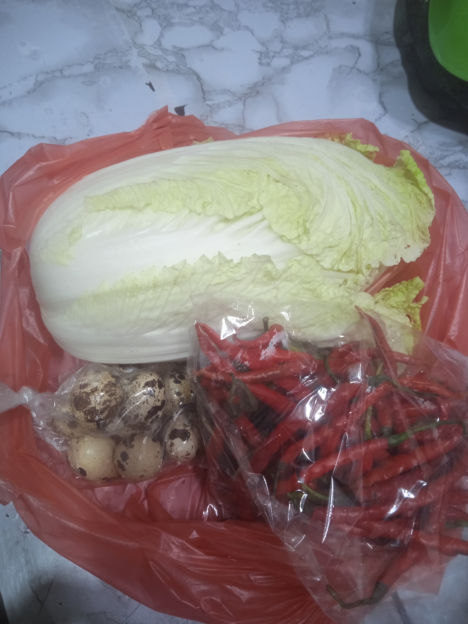 A large Chinese cabbage, a bag of red chilies, and several quail eggs are laid out on a red plastic bag against a marbled grey surface, possibly on a table or floor. The lighting suggests an indoor setting, likely in a residential kitchen. The scene appears to be a collection of fresh produce. There are no people or discernible actions taking place. The specific location within Surakarta, Indonesia, cannot be determined from the image alone.