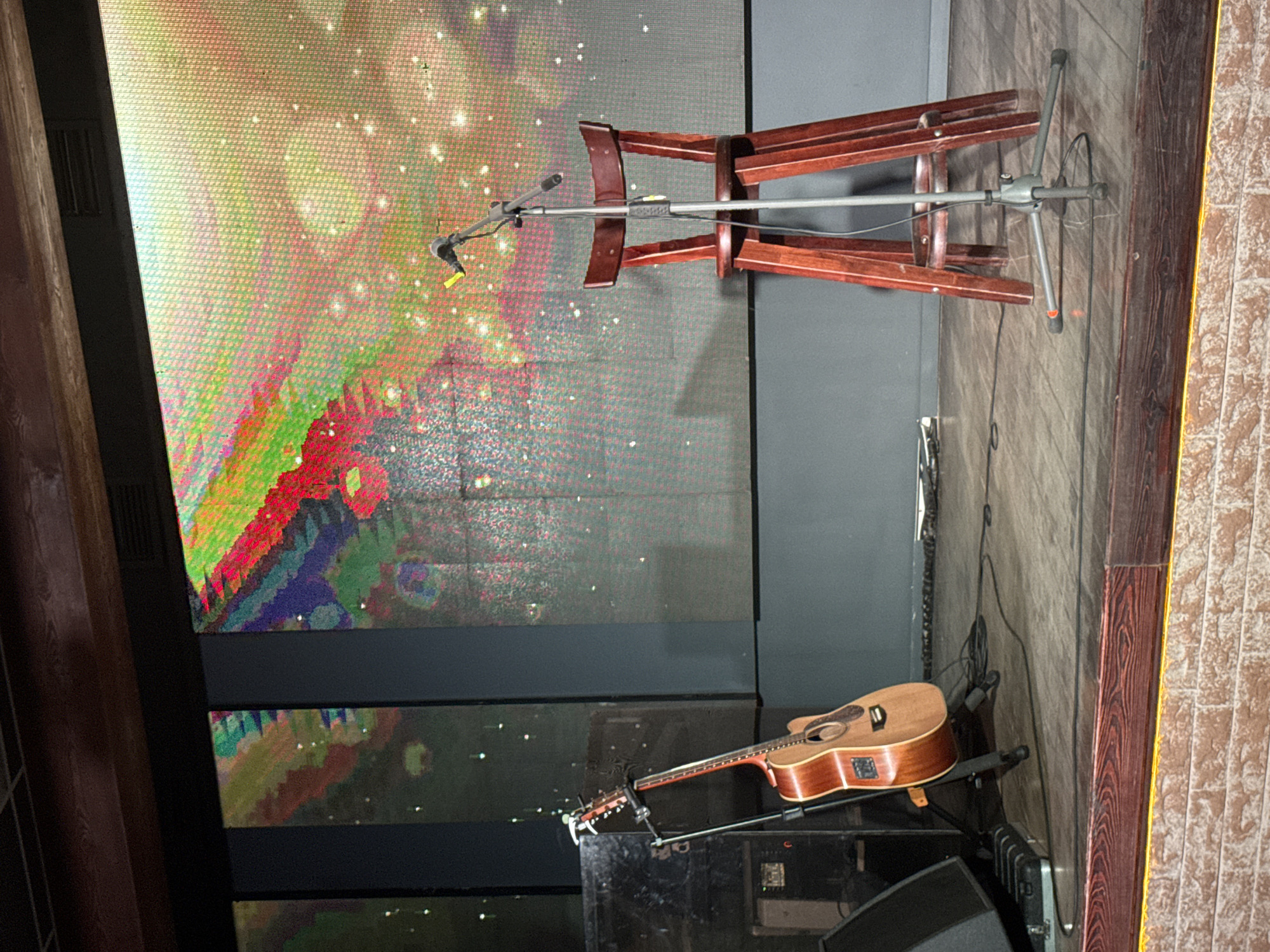 A tilted eye-level shot shows a stage set up for a performance. The main backdrop is a large screen displaying vibrant, abstract, pixelated art. In the foreground, a guitar rests on a stand, angled towards the right. At the top, a folding wooden chair rests upside down on a microphone stand. The stage is cluttered with equipment including speakers and cables. The right edge of the image shows a wall with wood paneling, possibly brick covered with wood, oriented vertically. Based on the fact that it is from Chaikovsky, Russia this is likely a stage being prepared for a musical performance or cultural event.