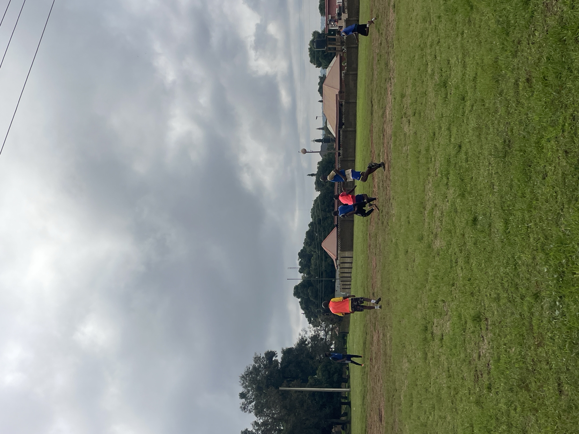 The image depicts a football practice session on a cloudy day, possibly in the city of Jos, Nigeria. Six individuals, dressed in athletic wear, are actively engaged in playing football on a grassy field. Two players are wearing bright orange vests over blue shirts, while the remaining four are in blue athletic attire. One player in an orange vest is positioned towards the left of the frame, facing away from the camera. Another player in an orange vest and patterned shorts is in the center, also facing away. To the right of the center, two players in blue are running. Further to the right, another player in blue is running. On the far left, a player in blue stands near a tall pole. The field is characterized by green grass with some patchy areas. In the background, there are trees, a fence, and residential buildings with red roofs, suggesting an urban or semi-urban setting. Overhead power lines are visible against the overcast sky.