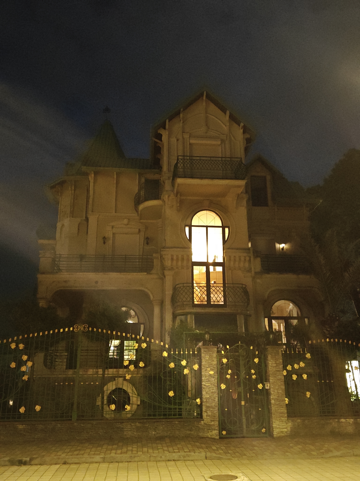 The image shows a large, ornate building behind a decorative fence at night. The building is several stories tall, with multiple balconies and a tower-like structure on one side. The architecture is elaborate, featuring arched windows and intricate detailing. Lights are visible inside the building, shining through the windows. The fence is made of metal and is adorned with yellow floral decorations. The foreground shows a paved area. The location is Unknown, Unknown.