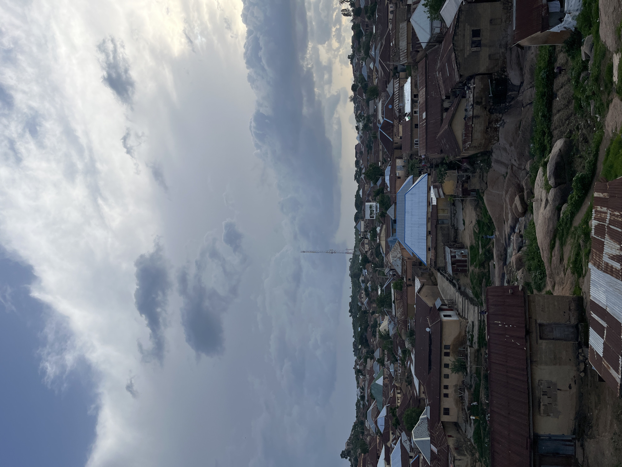 This is a high-angle, wide shot of a densely populated, rocky urban area in Jos, Nigeria. The scene is characterized by numerous houses with varied roof types, including corrugated metal and some with brightly colored metal roofs. The buildings appear to be constructed from earth-toned materials, suggesting a traditional architectural style. The terrain is uneven and rocky, with large boulders interspersed among the structures and vegetation. A tall communication tower is visible in the distance, above the houses. The sky is filled with dramatic, dark clouds, indicating an overcast or stormy condition. A few scattered figures are visible, appearing as small, indistinct shapes walking amongst the buildings, suggesting human presence and activity within the settlement.