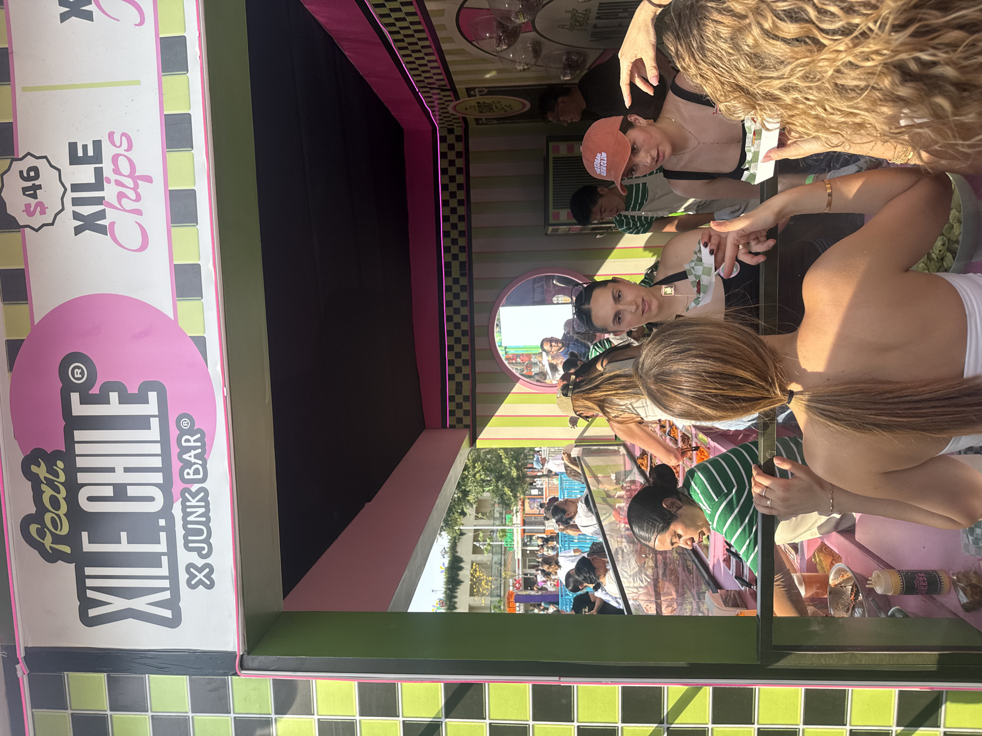 The image shows the XILE CHILE Junk Bar stand in Mexico City, Mexico. Several people are customers at the stand. They appear to be making purchases, while employees are preparing food. The stand's exterior has a green-and-black checkered pattern, with pink accents, and includes signage indicating the price of "XILE Chips" as $46. A mirrored surface on the stand reflects the crowd of people outside the enclosure.