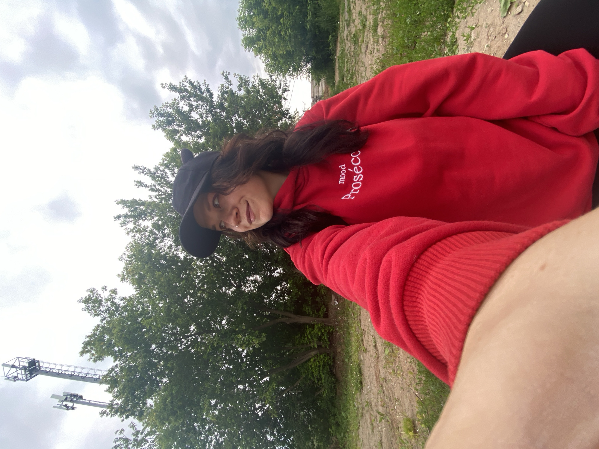 A young woman wearing a red hoodie and a black baseball cap is taking a selfie outdoors. The hoodie has white text that says "mood Prosecco." She has brown hair and is looking off to the left of the frame with a slight smile. Behind her are trees and two tall communication towers. The ground is dirt and grass. The sky is overcast. The image appears to have been taken near the city of Chaikovsky, Russia, given the presence of communication towers and the general landscape.