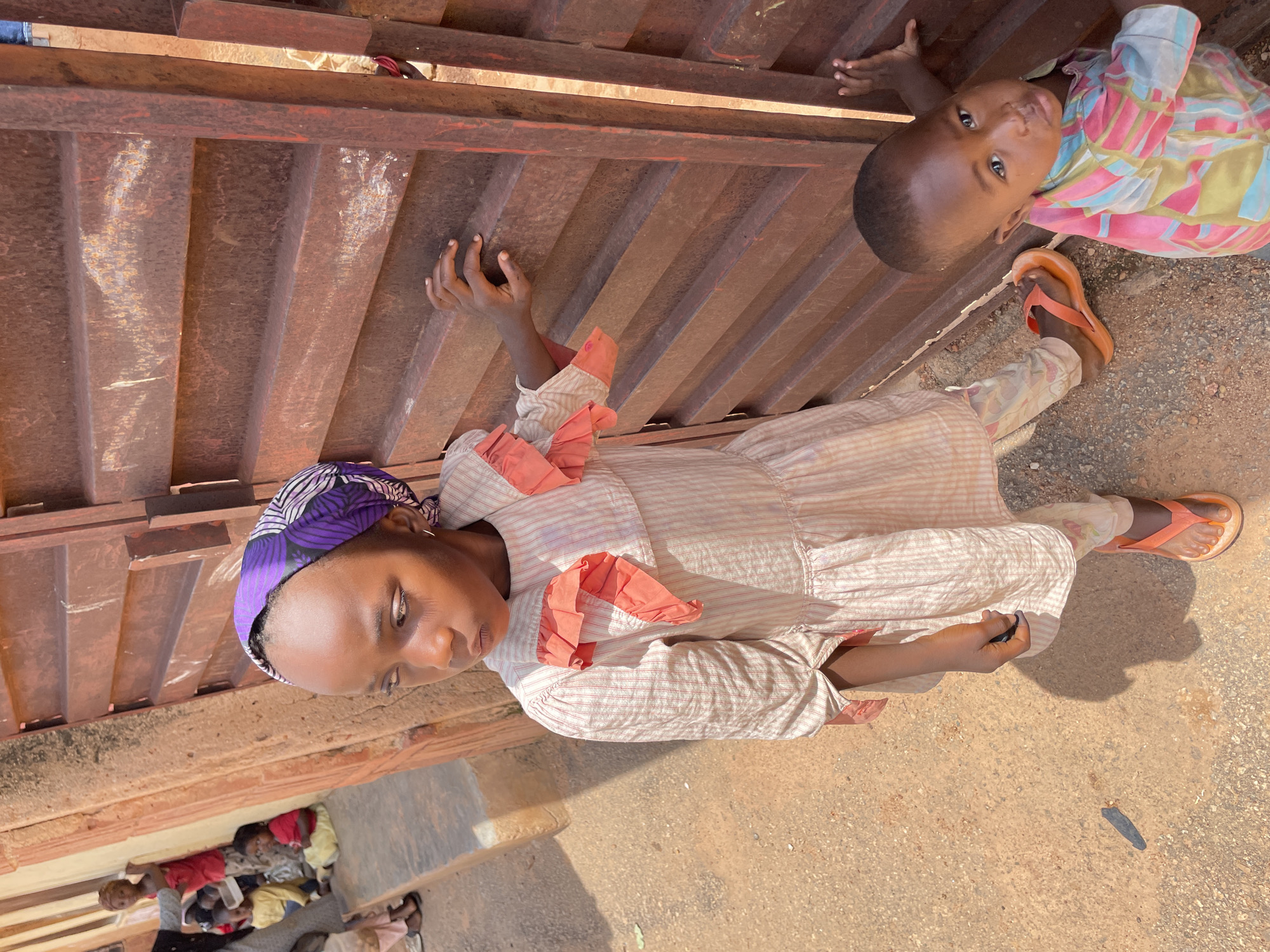 In Jos, Nigeria, a young girl stands in front of a rusted metal gate, her head adorned with a purple and white patterned headscarf. She wears a cream-colored dress with subtle vertical stripes and coral-colored ruffles around the collar and cuffs. Her legs are clad in patterned leggings, and she sports orange flip-flops. To her right, a toddler in a brightly colored, striped shirt leans against the gate, looking towards the camera. The gate, made of horizontal metal bars, shows signs of rust and wear. In the background, blurred figures of other children are visible, some sitting on a ledge. The ground is a mixture of dirt and gravel, with harsh sunlight casting distinct shadows.