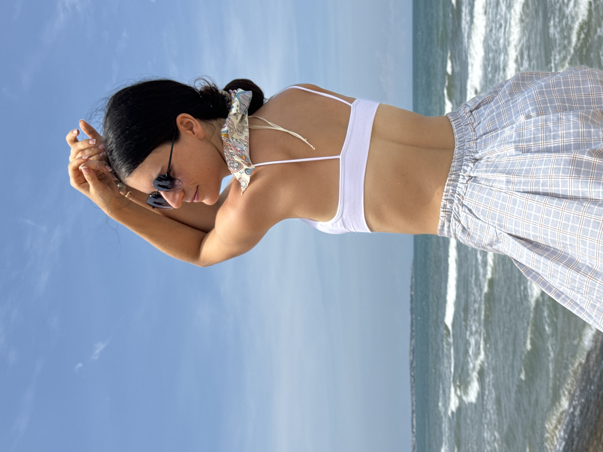 A woman stands on a rocky outcrop overlooking the ocean in Mũi Né, Vietnam. She is wearing a white bikini top, a light blue and white checkered skirt, and sunglasses. Her arms are raised above her head, and a patterned bandana is tied around her neck. The ocean has moderate waves, and the sky is clear with a few wispy clouds.