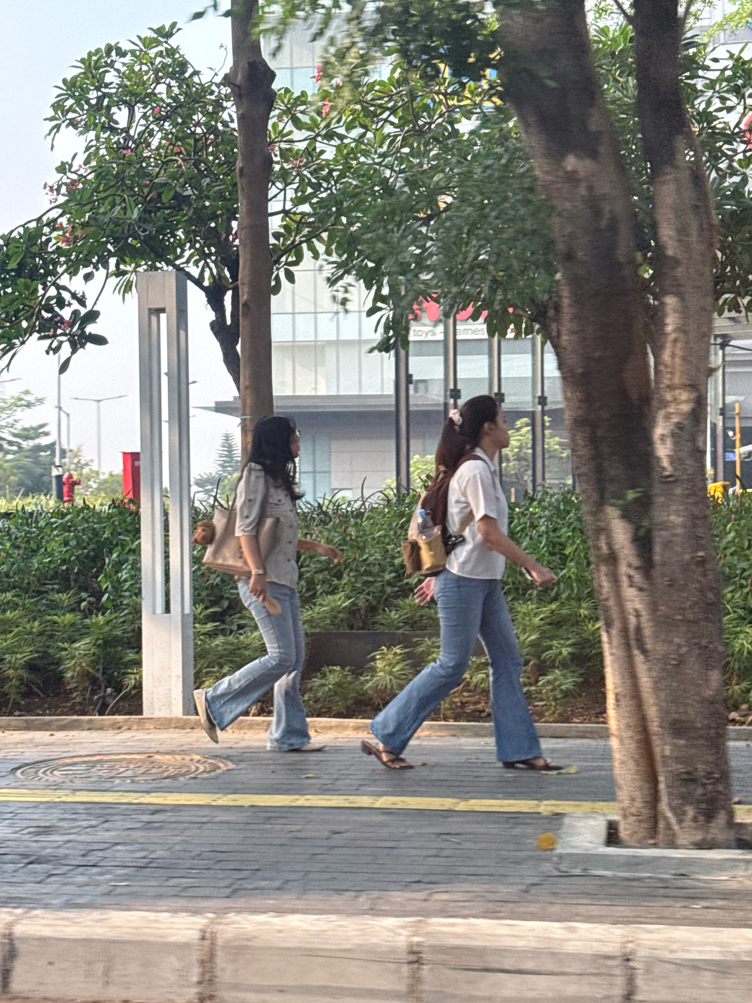 In Surabaya, Indonesia, two women are walking on a paved sidewalk adjacent to a road. The woman on the left, wearing a light-colored, collared shirt and flared jeans, carries a large tote bag on her right shoulder. The woman on the right, in a white collared shirt and flared jeans, has a backpack with a water bottle attached. Both women are walking towards the right of the frame. Behind them, lush green bushes form a dense hedge, and further back, a modern building with a glass facade is visible. Trees with dark bark and green foliage are interspersed throughout the scene, with one prominently positioned in the foreground to the right, partially obscuring the background. A street light pole stands to the left of the women, and a fire hydrant is visible behind the bushes. The ground is a patterned brick walkway with a yellow stripe marking the edge of the pedestrian area.