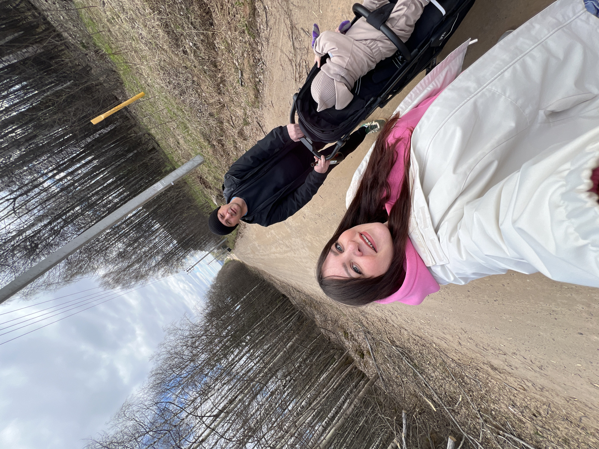 A family—two adults and a child in a stroller—are walking along a sandy path flanked by birch trees. The path is in a linear clearing through a birch forest. The stroller is black, and the child is wearing a beige outfit. The adults are dressed in outerwear appropriate for cool weather; the adult in the foreground is wearing a white coat with a pink hooded sweatshirt underneath, and the other is wearing a black jacket and beanie. A utility pole and power lines are visible in the background, extending towards the horizon. Based on the birch trees and utility infrastructure, the location is likely Noviy, Russia.