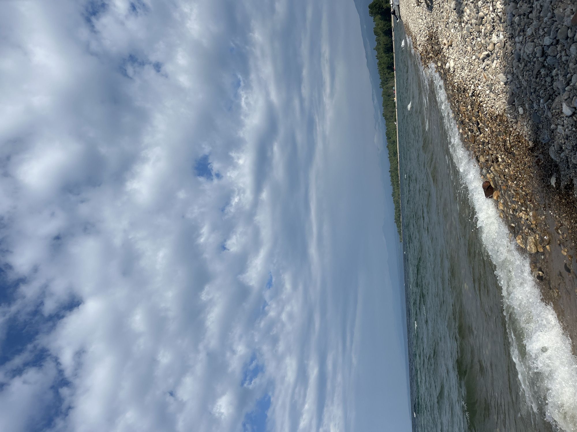 The image shows a wide, pebble-strewn beach along a large body of water. The water is a dark, choppy green with white-capped waves rolling onto the shore. The sky is filled with a dramatic arrangement of grey and white clouds, with patches of blue sky visible between them. In the distance, the shoreline is lined with a dense forest of green trees, beyond which faint, hazy mountains can be seen. A few scattered tents and vehicles are visible along the tree line. The overall impression is of a natural, somewhat wild landscape. The exact location in Tolbazikha, Russia, is not specified in the visual information.