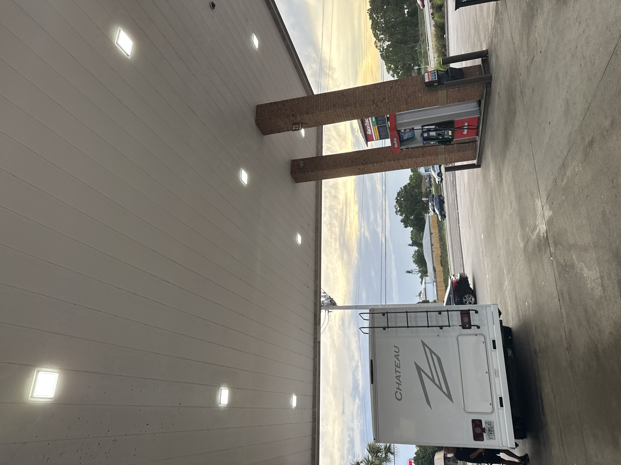 A white Chateau RV is parked at a RaceTrac gas station in Palm Bay, Florida. The back of the RV, including its ladder, is visible. A man is standing behind the RV. A car is visible in the background. The gas station has brick pillars and a price display. The sky is visible in the background.