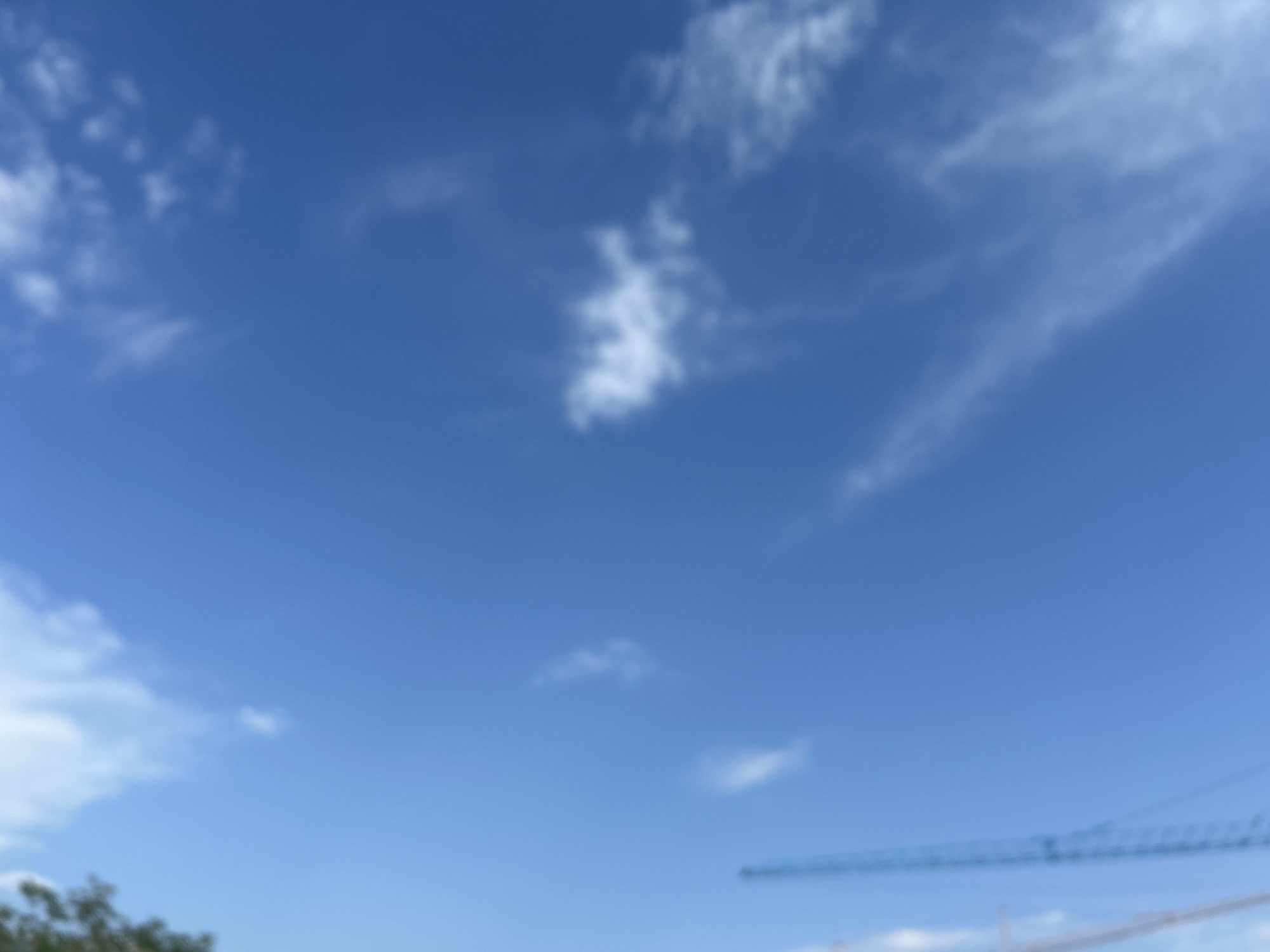This media file is a slightly blurred photograph of a clear blue sky with sparse white clouds. In the bottom right corner, a portion of a blue construction crane is visible, along with some very faint white clouds near the horizon. The top left of the image shows the blurred green foliage of trees. The lighting suggests it is daytime, and the clear sky indicates fair weather. The presence of the construction crane hints at an ongoing building project, possibly in an urban or developing area. The overall mood is serene and expansive, due to the focus on the sky. No text is visible in the image. The location context provided is Boreti, Montenegro.