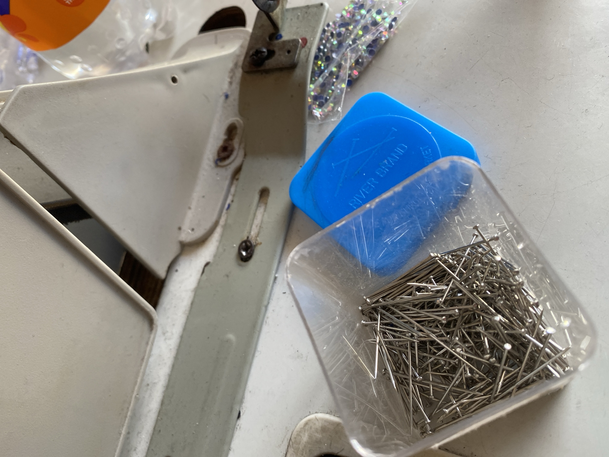 The image shows a close-up, overhead view of a white sewing machine and various sewing supplies on a white surface. In the foreground, a clear plastic container is filled with numerous silver sewing pins. To the right of the pins is a bright blue, circular object with the words "RIVER BRAND" embossed on it. Further to the right, a small clear plastic bag contains an assortment of small, colorful rhinestones or sequins. The sewing machine appears to be made of metal and plastic and shows some signs of wear. The background is out of focus but suggests an indoor environment. There are no people visible in the image. The scene is consistent with a workspace for sewing or crafting. There is no specific information in the image that would allow for the identification of its location within Jalingo, Nigeria.