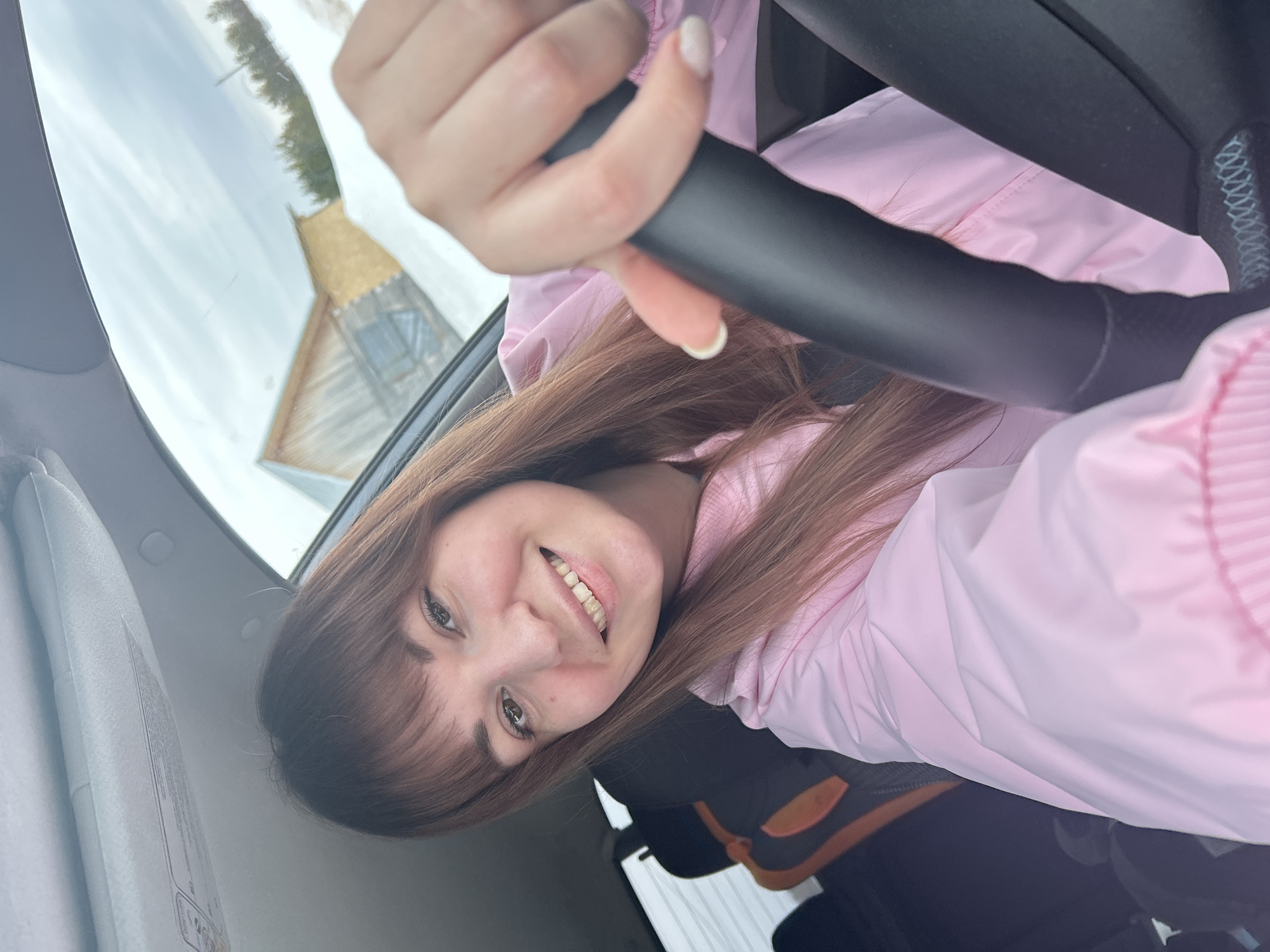 A smiling woman in a pink jacket sits in a car, holding the steering wheel. Through the windshield, a wooden structure with a straw roof is visible. The scene appears to be in Фоки, Russia.