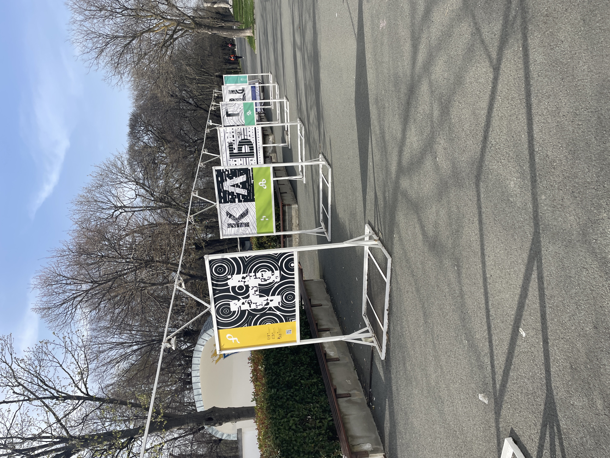 A series of white metal frames display poster-like signs in a row along a paved road. The signs feature graphic designs in black and white, some incorporating elements of green and yellow. Bare trees line the edge of the road, and the sky is visible in the background with patches of blue. The road surface appears asphalt or similar material.
The first sign, in the lower foreground, has a black and white circular pattern on the upper half and a yellow section on the lower half, featuring smaller graphics. This sign appears to be situated adjacent to a low wall and a hedge. The signs further down the line show distinct letter-like forms against green or white backgrounds.
The location is in the city of Burgas, Bulgaria.