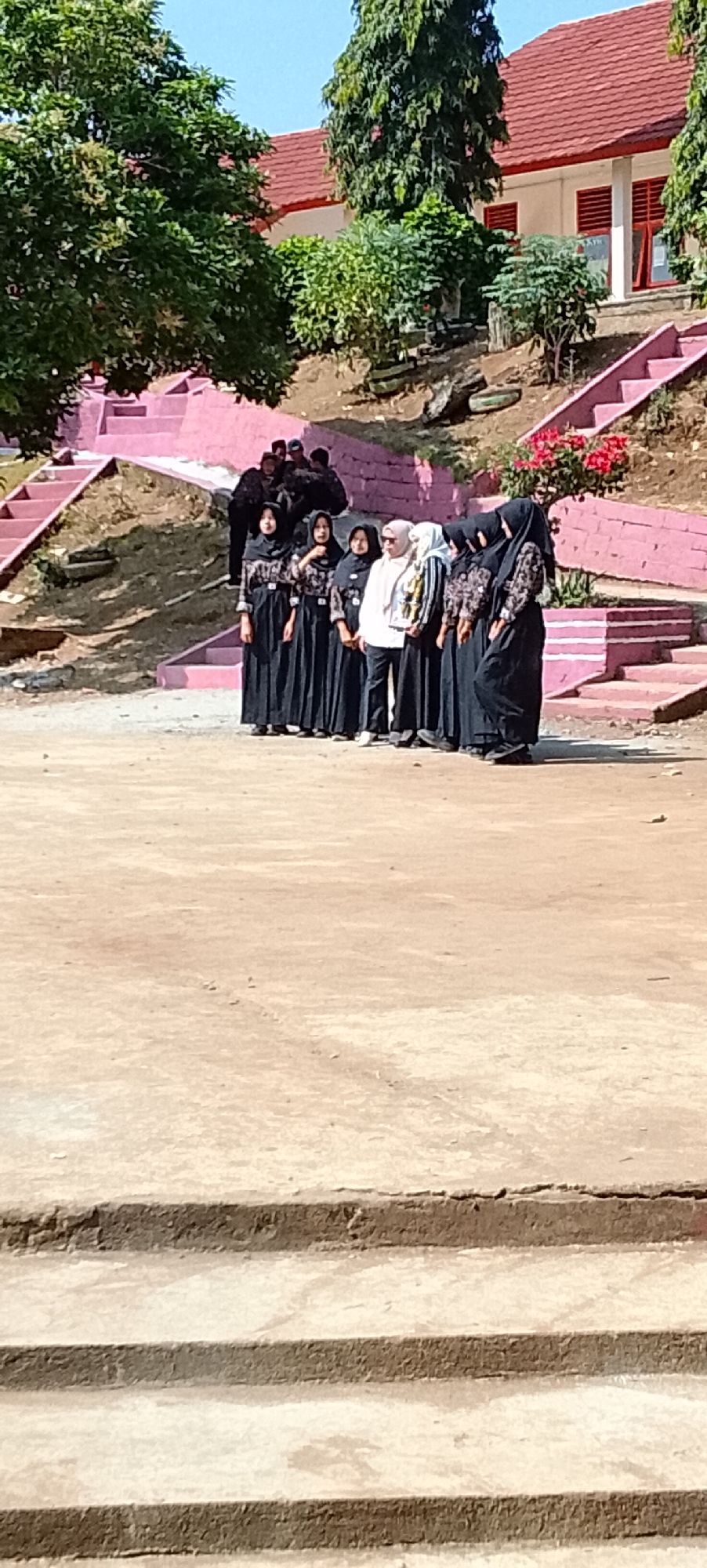 The image shows a group of people, predominantly women wearing hijabs and dark, patterned clothing, standing in what appears to be an outdoor school or public space. They are arranged in a line, facing slightly to the right. Behind them, there are pink stairs leading up to a building with a red roof. Trees and bushes are also visible in the background. The ground in front of the group is a dirt or concrete surface. The scene suggests a formal gathering or event taking place during daylight. The location is identified as Jepara, Indonesia, an area known for its educational institutions and public spaces.